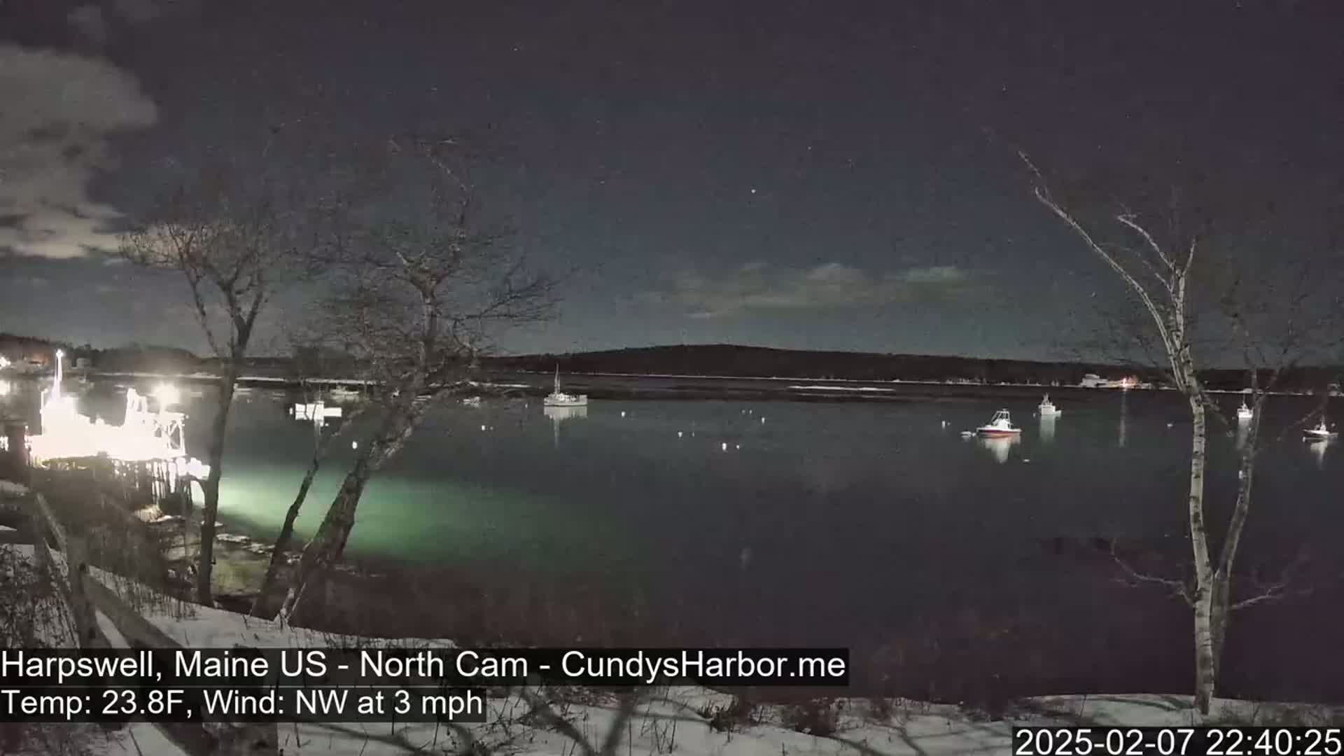 A nighttime view of a calm harbor with several boats, under a partly cloudy sky, illuminated by lights on a nearby dock.