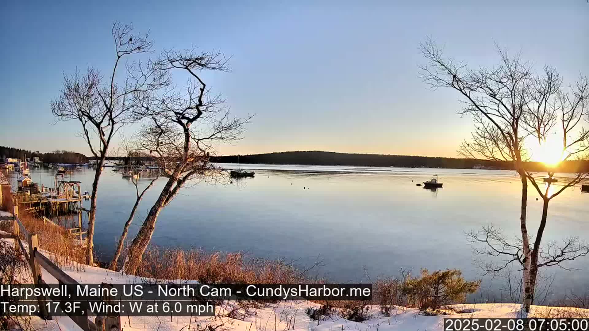 A calm, partially frozen harbor at sunrise is viewed from a snow-covered shoreline, with bare trees in the foreground and several boats scattered across the water.
