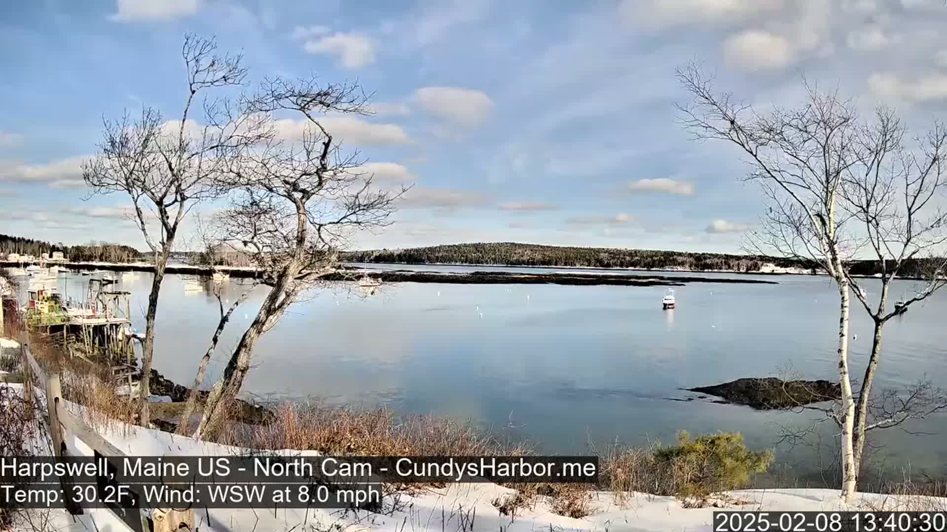A partly cloudy, cold day shows a calm harbor with several boats, viewed from a snow-covered shoreline with bare trees.