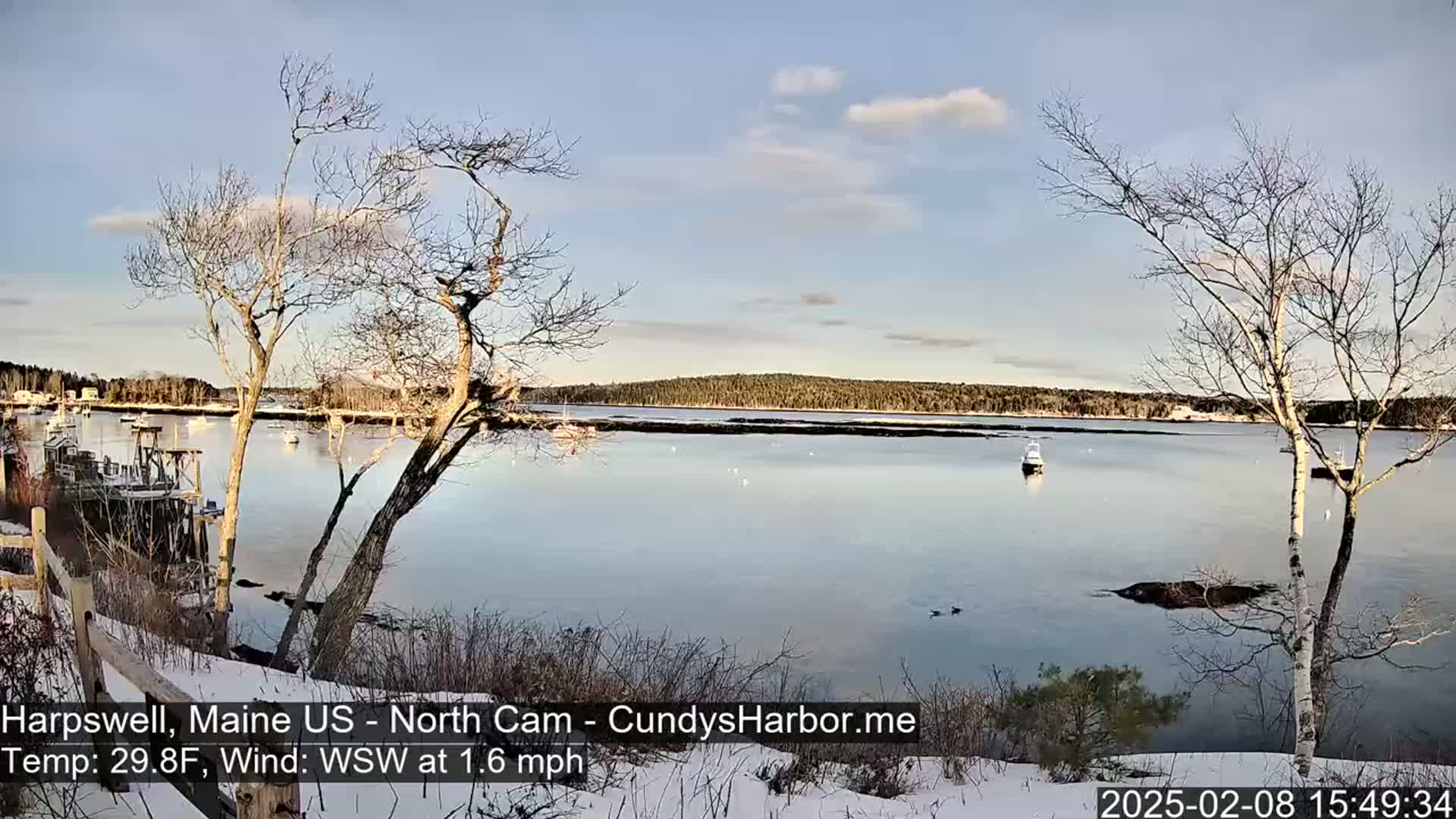 A calm, partly cloudy day overlooks a snow-dusted harbor with several boats, trees without leaves, and a shoreline.