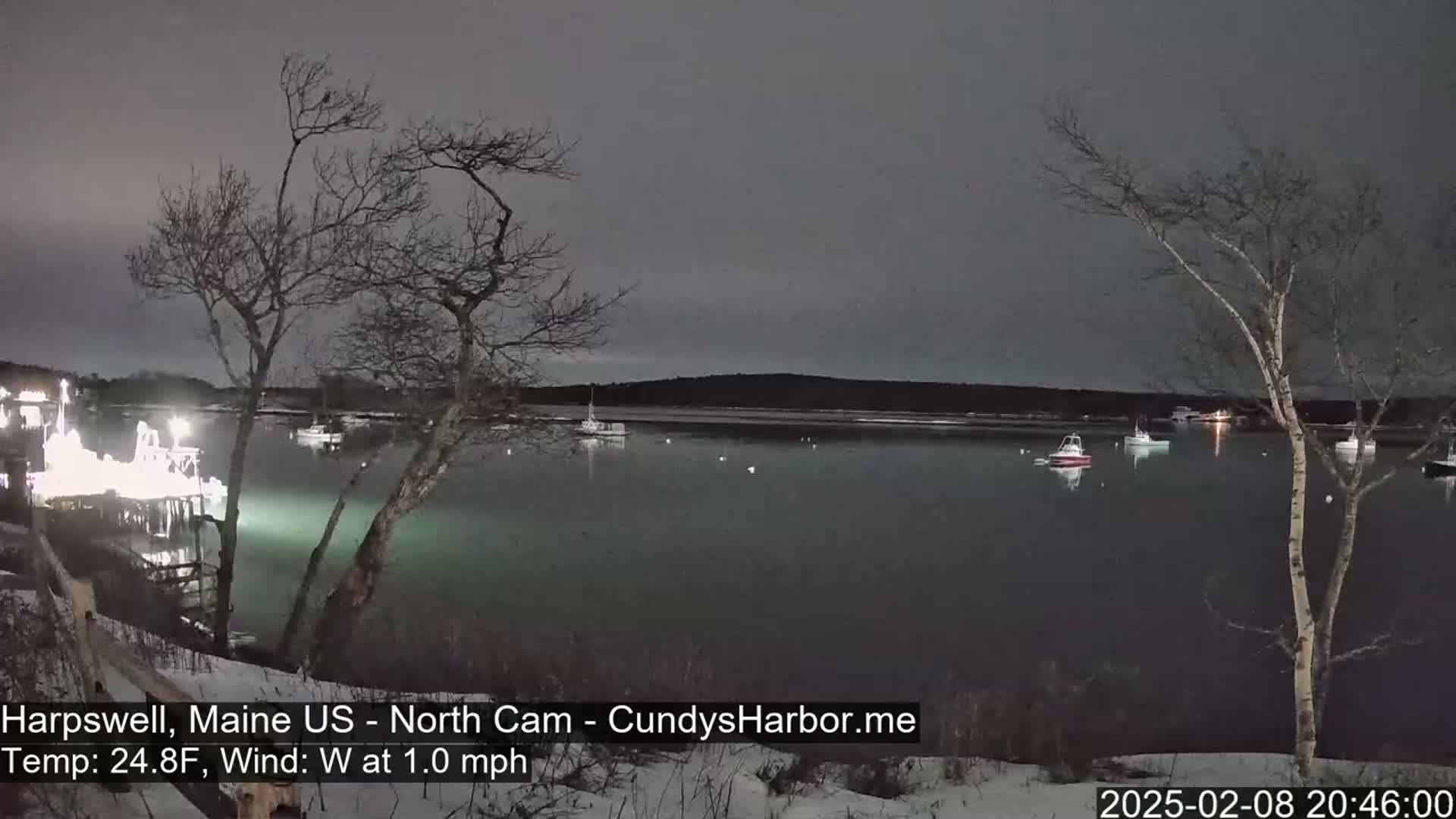 A nighttime view of a calm harbor with several boats at anchor, under a dark, overcast sky, with a light dusting of snow on the ground.