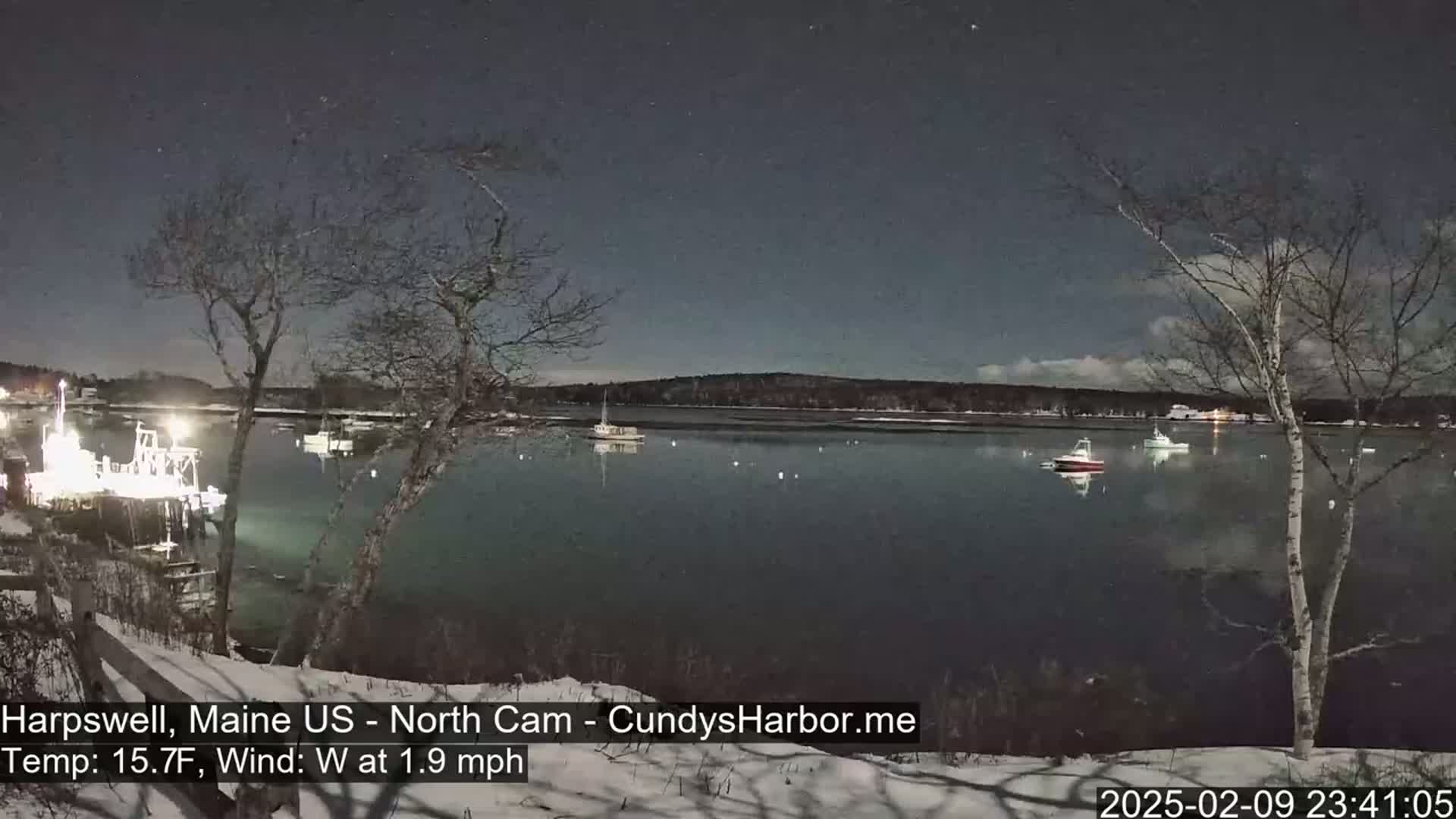 Under a dark, starry sky, several boats are moored in a calm, dark-water harbor, partially covered in snow and surrounded by leafless trees.