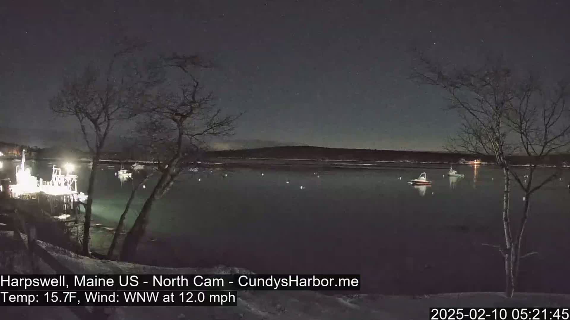 A nighttime view of a calm body of water with several boats, viewed from a snow-covered shoreline under a starry sky with a 12 mph wind.