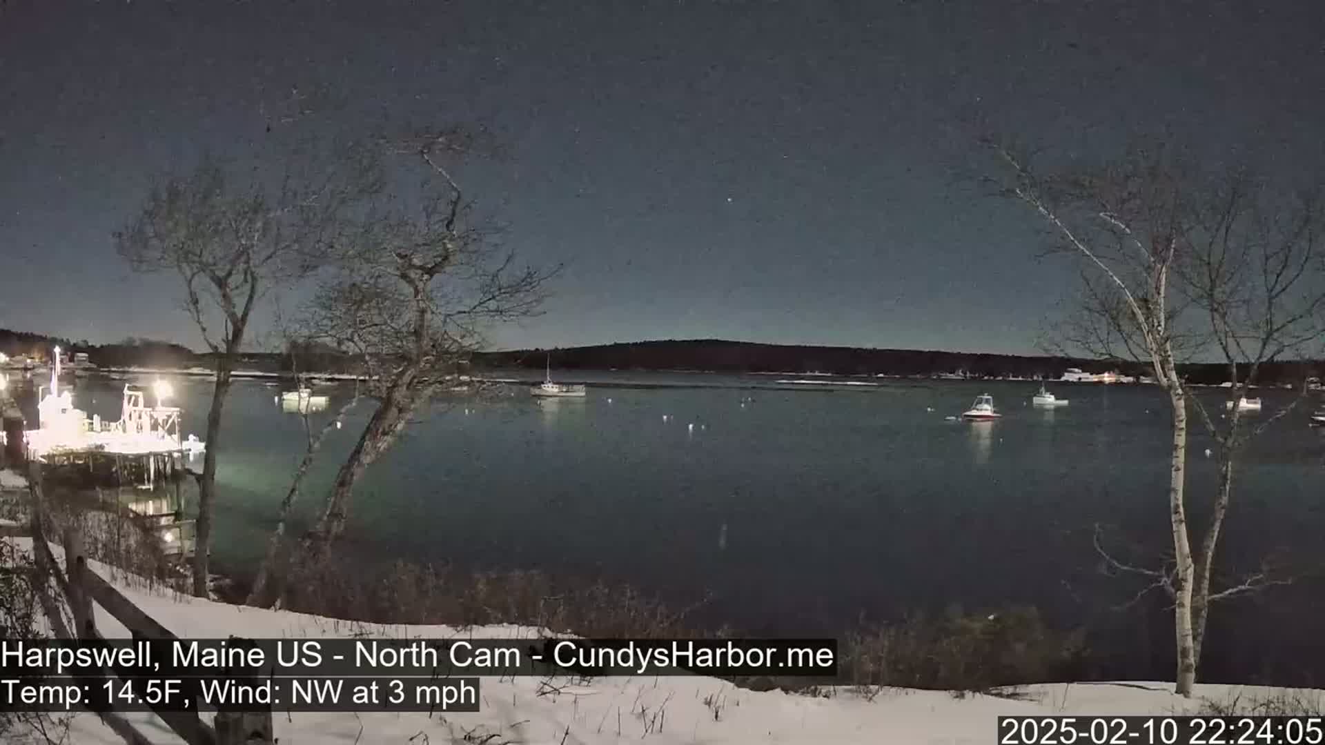 A nighttime view of a harbor with several boats moored, under a clear, dark sky, with snow on the ground and a light northwest wind.