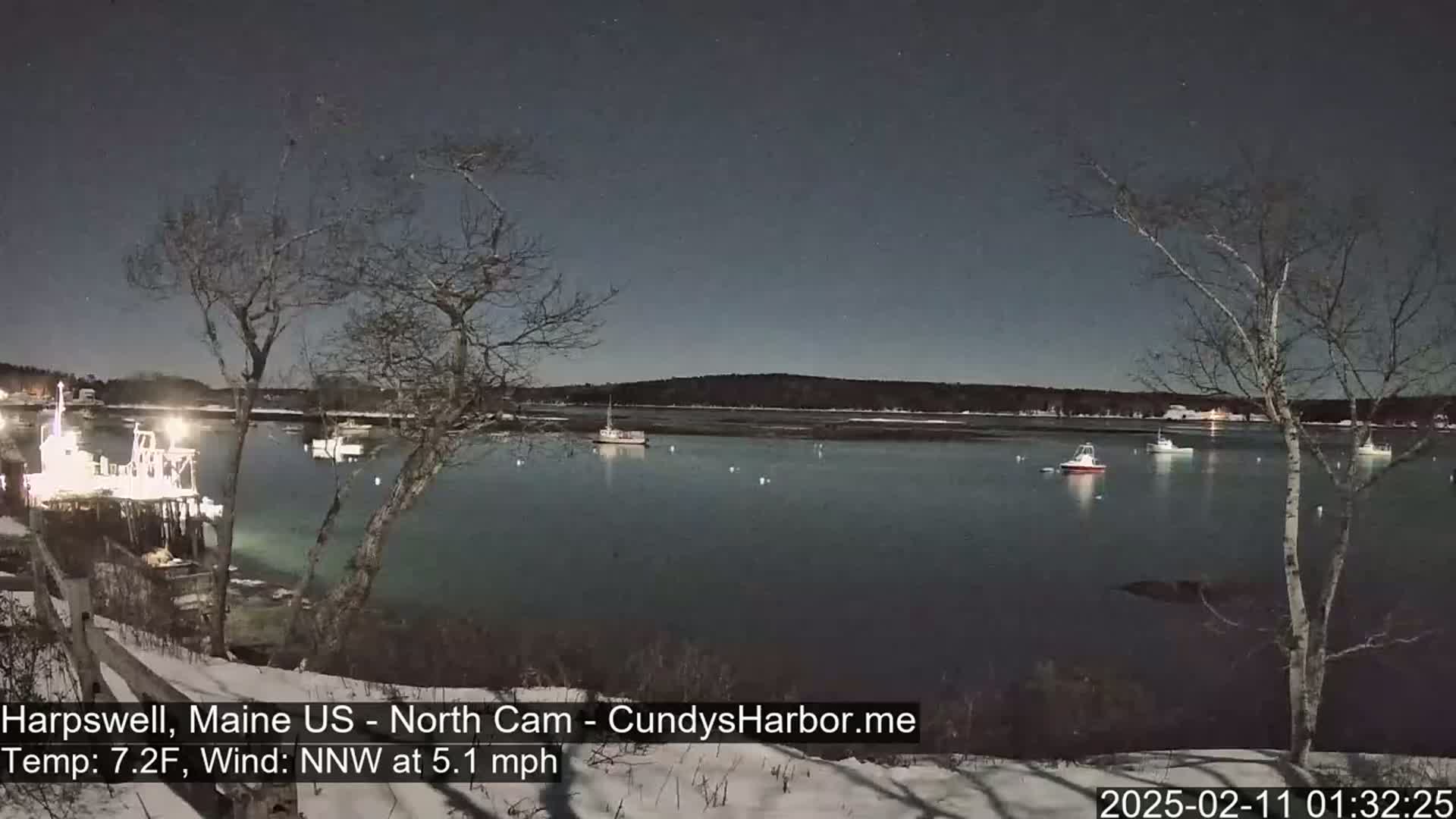 A moonlit harbor at night, with several boats moored near a snow-covered shoreline under a clear, dark sky and a light northwest wind.