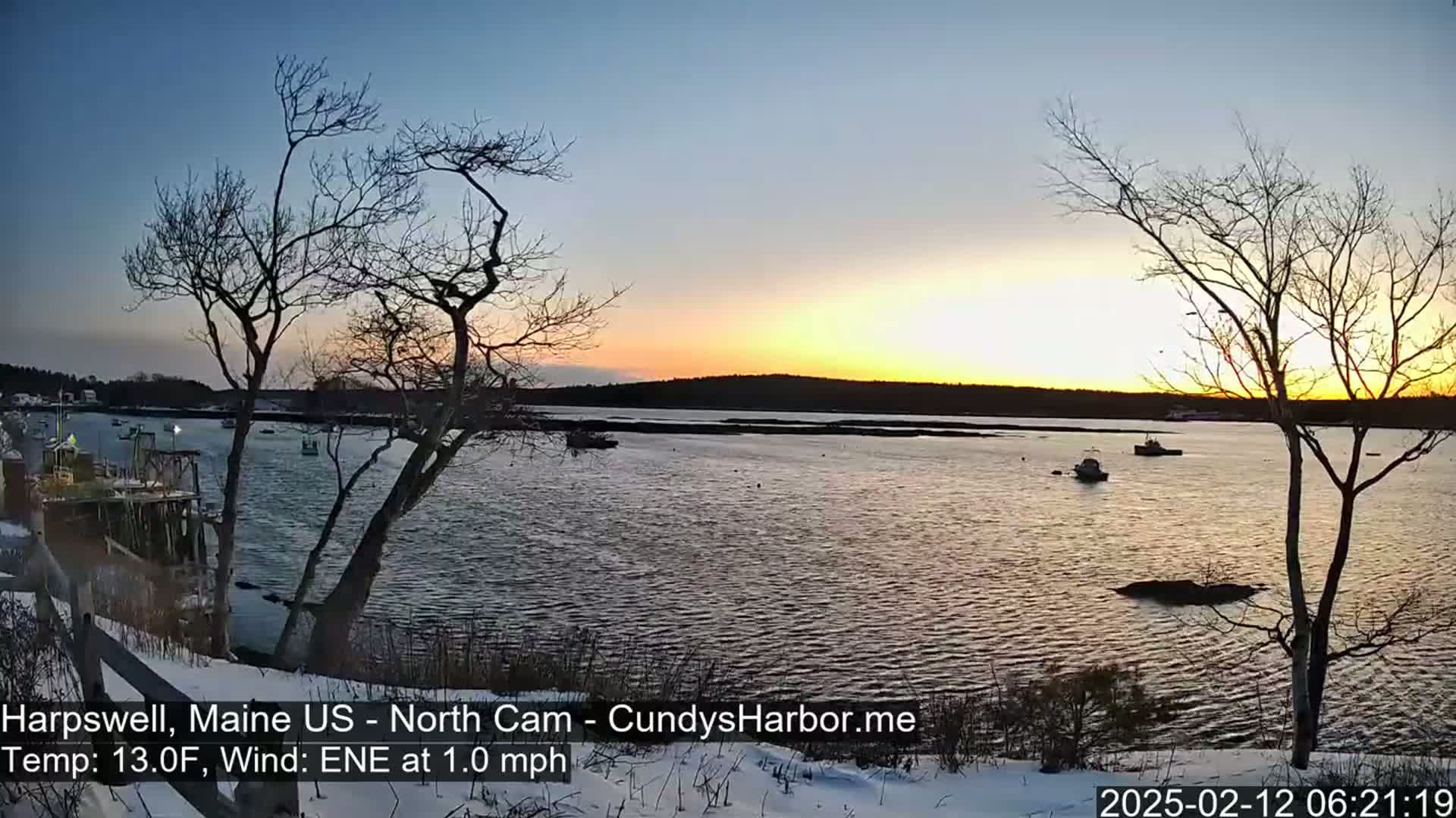 A calm body of water at sunset, viewed from a snow-covered shoreline with bare trees, under a clear sky.