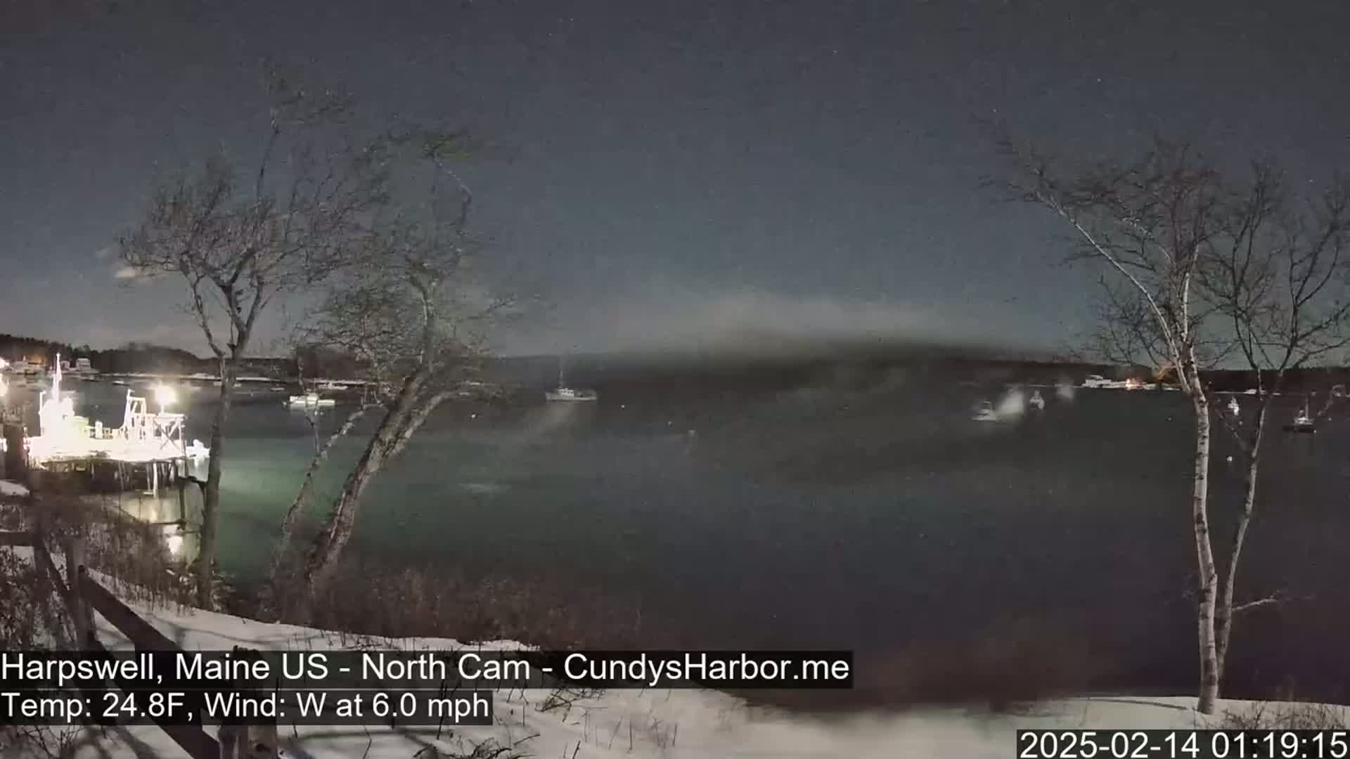 A nighttime view of a snow-covered harbor with boats and a dock under a mostly clear, dark sky, with a 6.0 mph westerly wind and a temperature of 24.8°F.