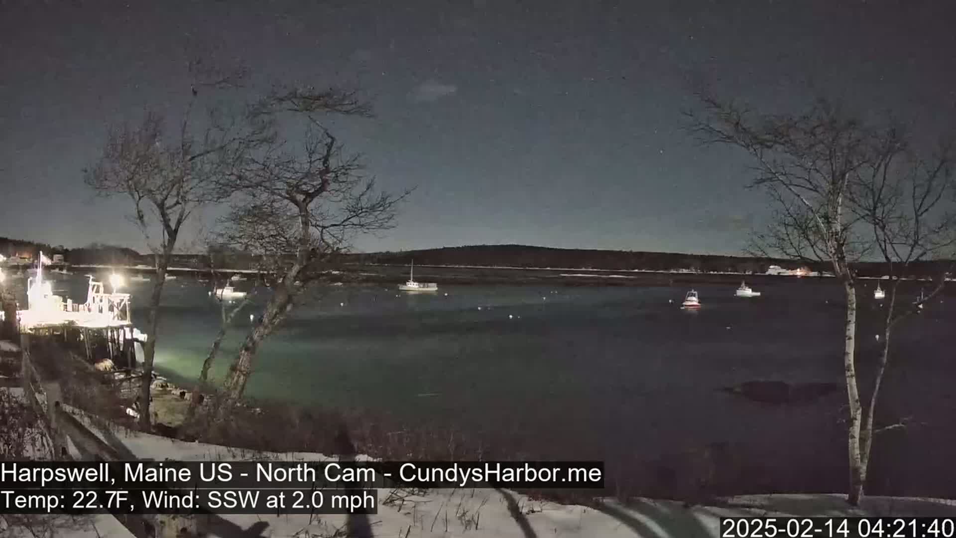 A dark, calm harbor at night, under a mostly clear sky, with several boats moored and a few lights visible on the shore, a light snow cover on the ground and a slight south-southwesterly wind.