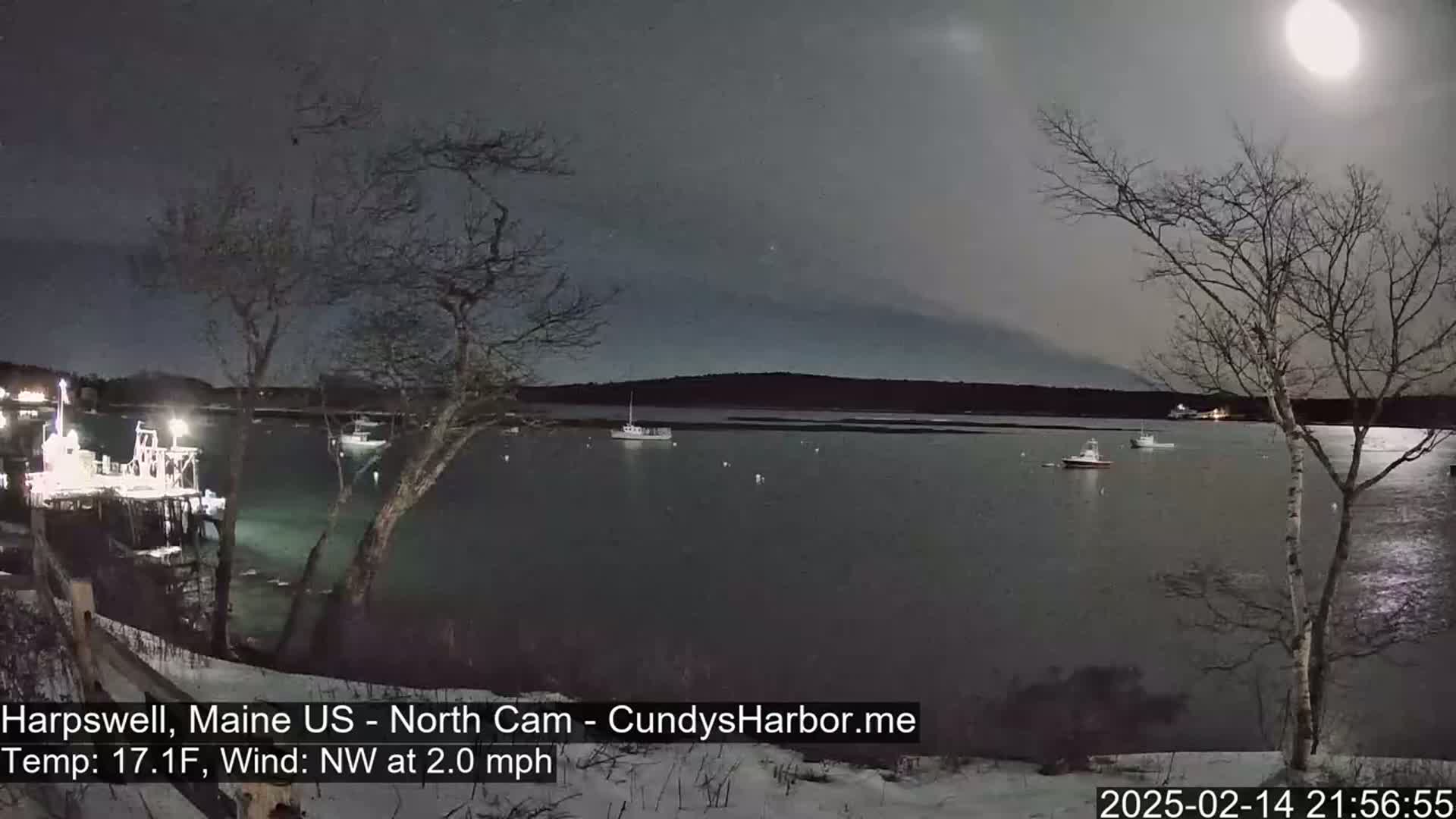 Under a bright moon and a mostly cloudy night sky, several boats are moored in a calm harbor, viewed from a snowy shoreline with bare trees.
