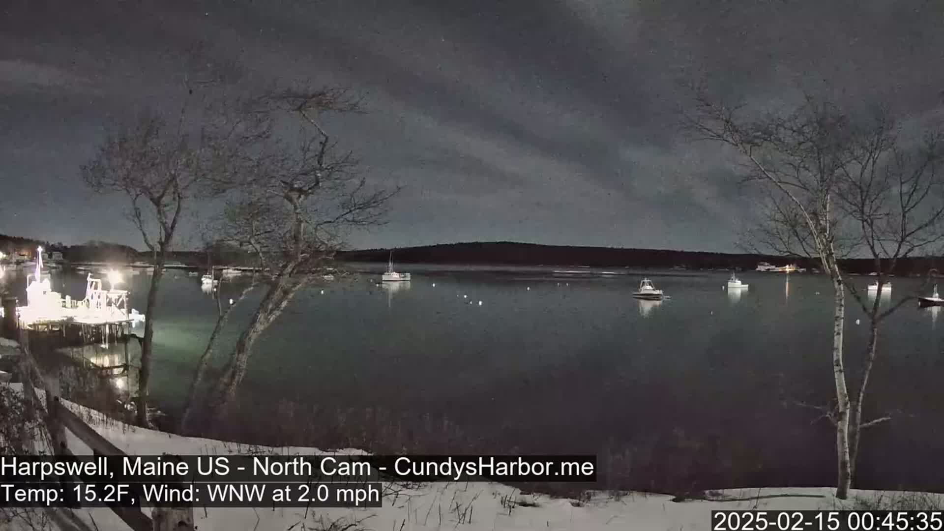 A nighttime view of a calm harbor with several boats moored, under a dark, cloudy sky, with snow on the ground and a light wind.