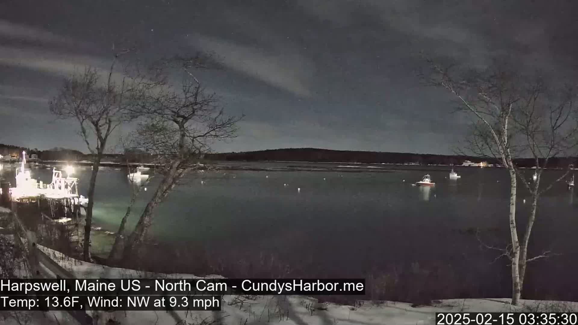 A nighttime view of a calm harbor with several boats moored, under a partly cloudy sky, with snow on the ground and a northwest wind.