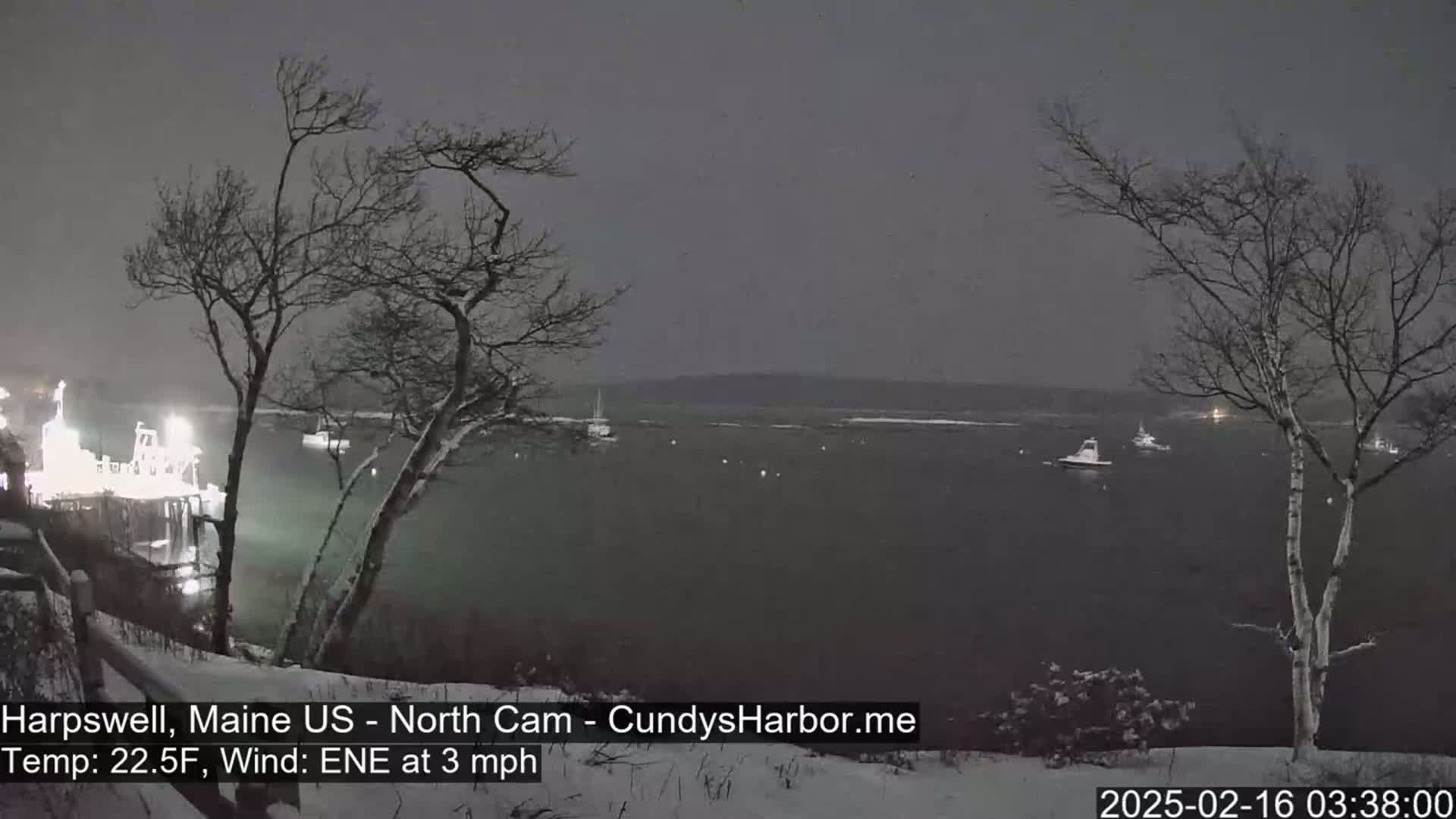 A snow-covered landscape at night shows a dark body of water with several boats, a lit structure on the shore, and bare trees dusted with snow.