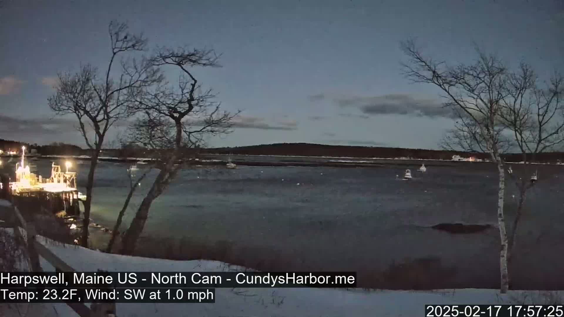 A dark, calm body of water with several boats is seen from a snowy shoreline under a twilight sky.