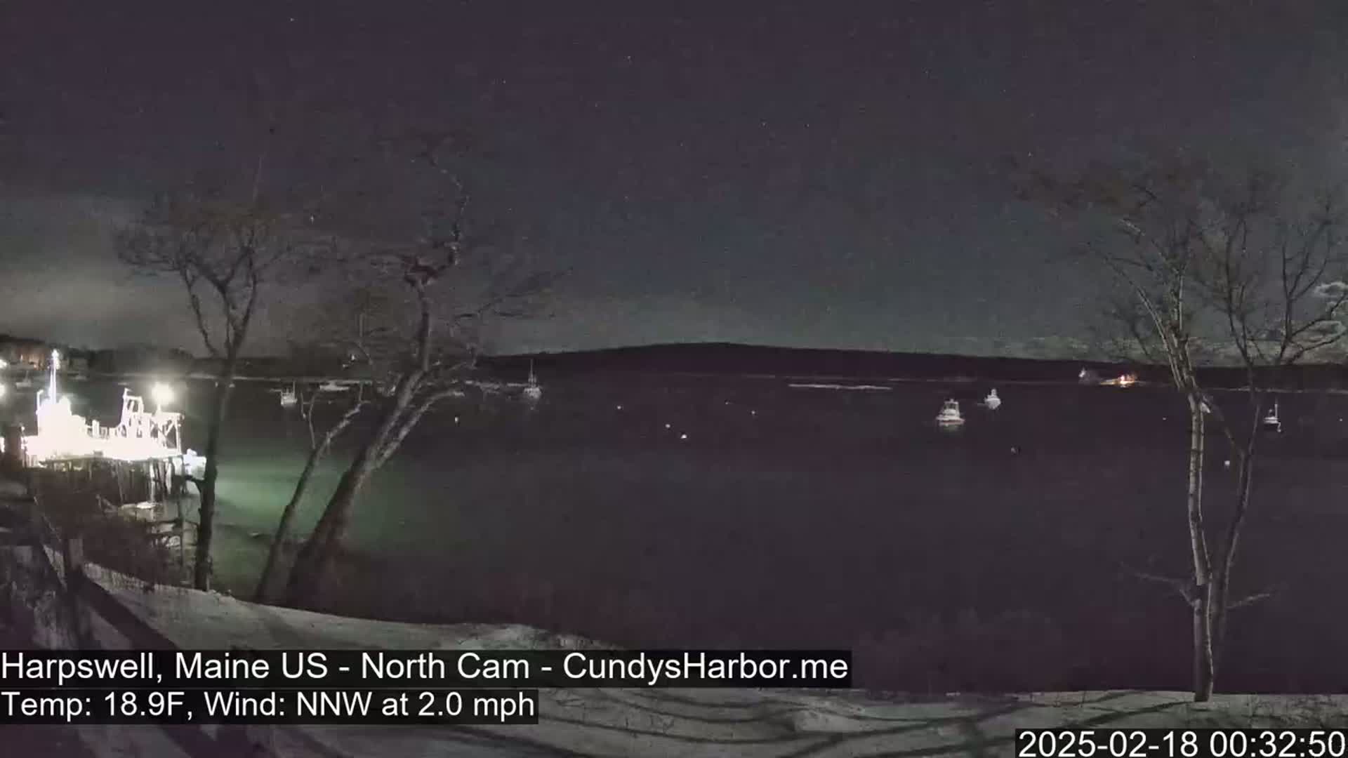 A nighttime view of a harbor with several boats, under a clear, starry sky, with a light snow cover on the ground and a slight wind.