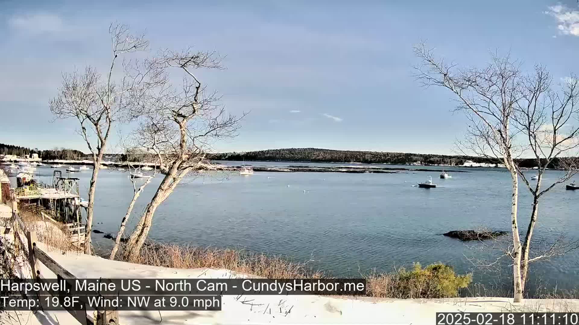A calm, clear day shows a snow-dusted shoreline overlooking a bay with several boats, under a mostly clear blue sky.