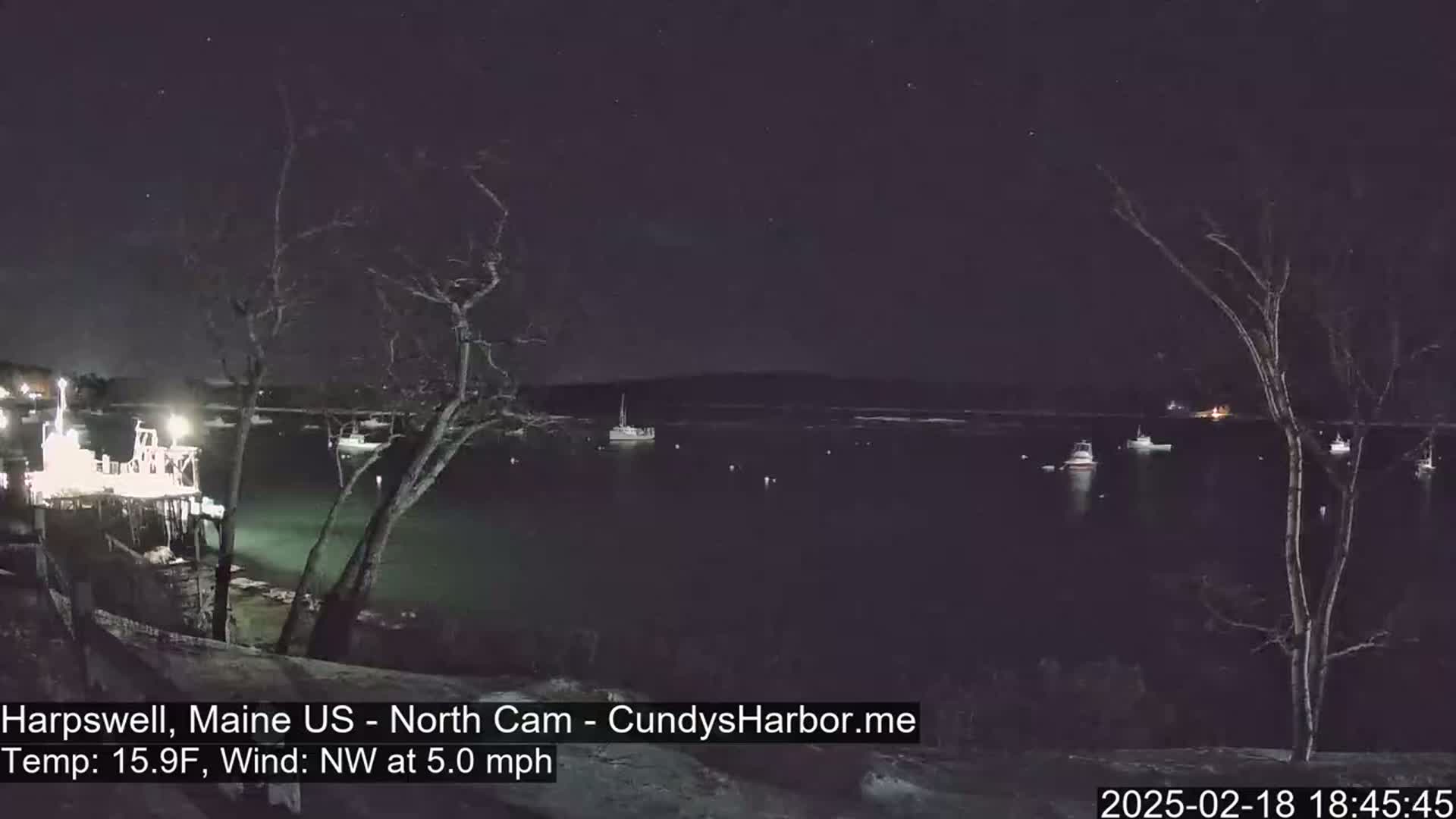 A nighttime view of a harbor with several boats, under a clear, starry sky, with a northwest wind at 5 mph and a temperature of 15.9°F.