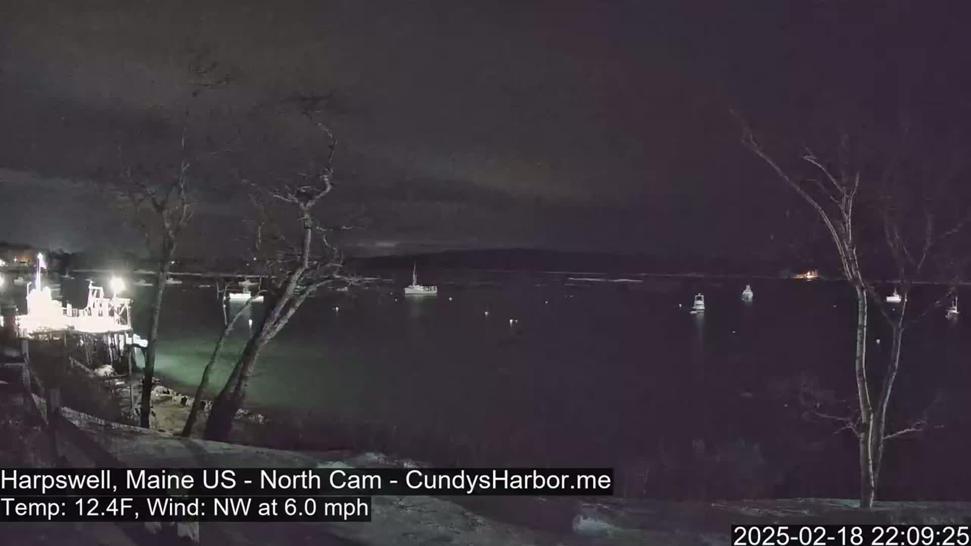 A nighttime view of a calm body of water with several boats and a pier, under a dark, cloudy sky with a northwest wind.