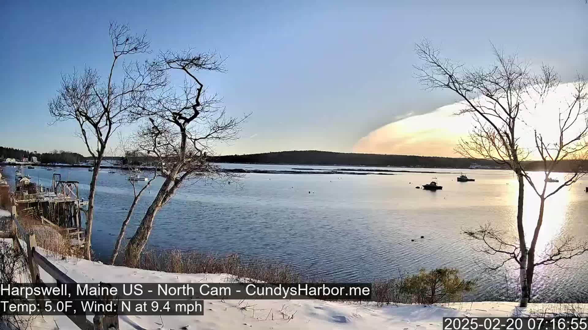 A calm bay with several boats is viewed from a snow-covered shoreline under a partly cloudy, pale-orange sunset, with bare trees in the foreground and a light north wind.