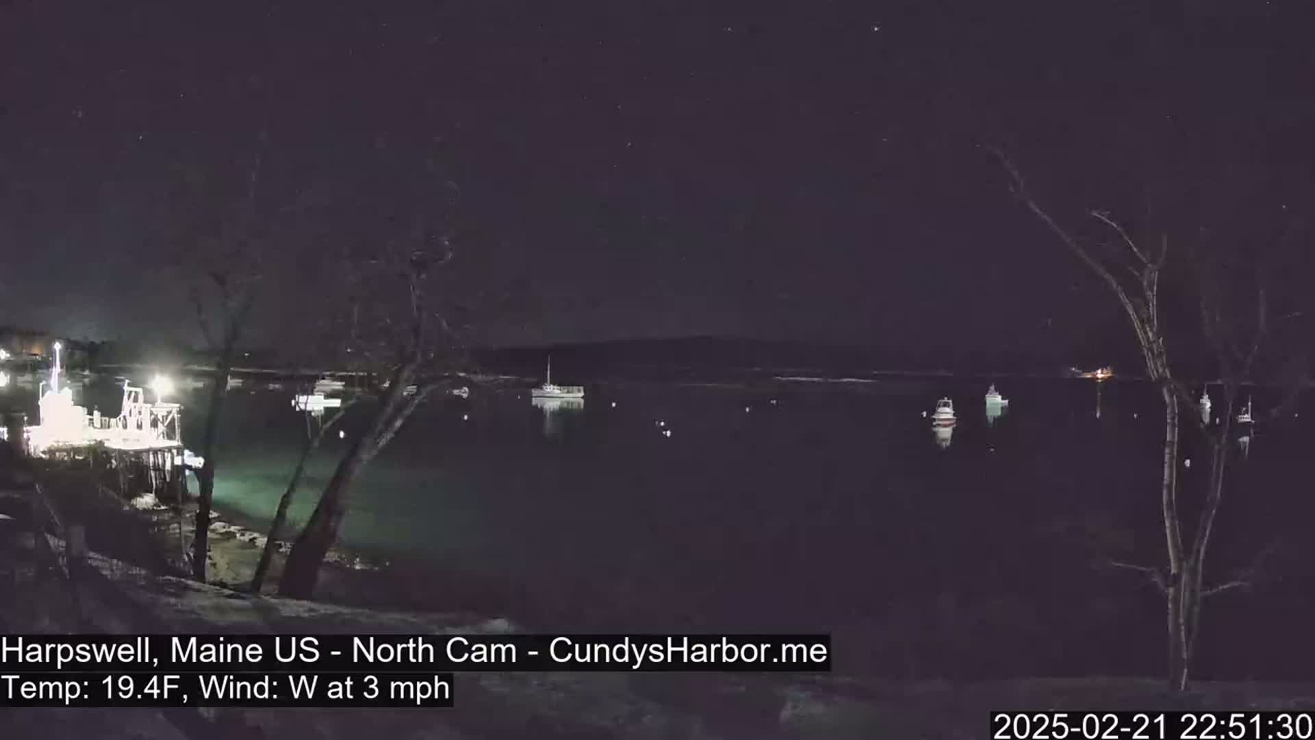 A nighttime view of a calm body of water with several boats moored, under a starry sky, and a slight westerly wind.