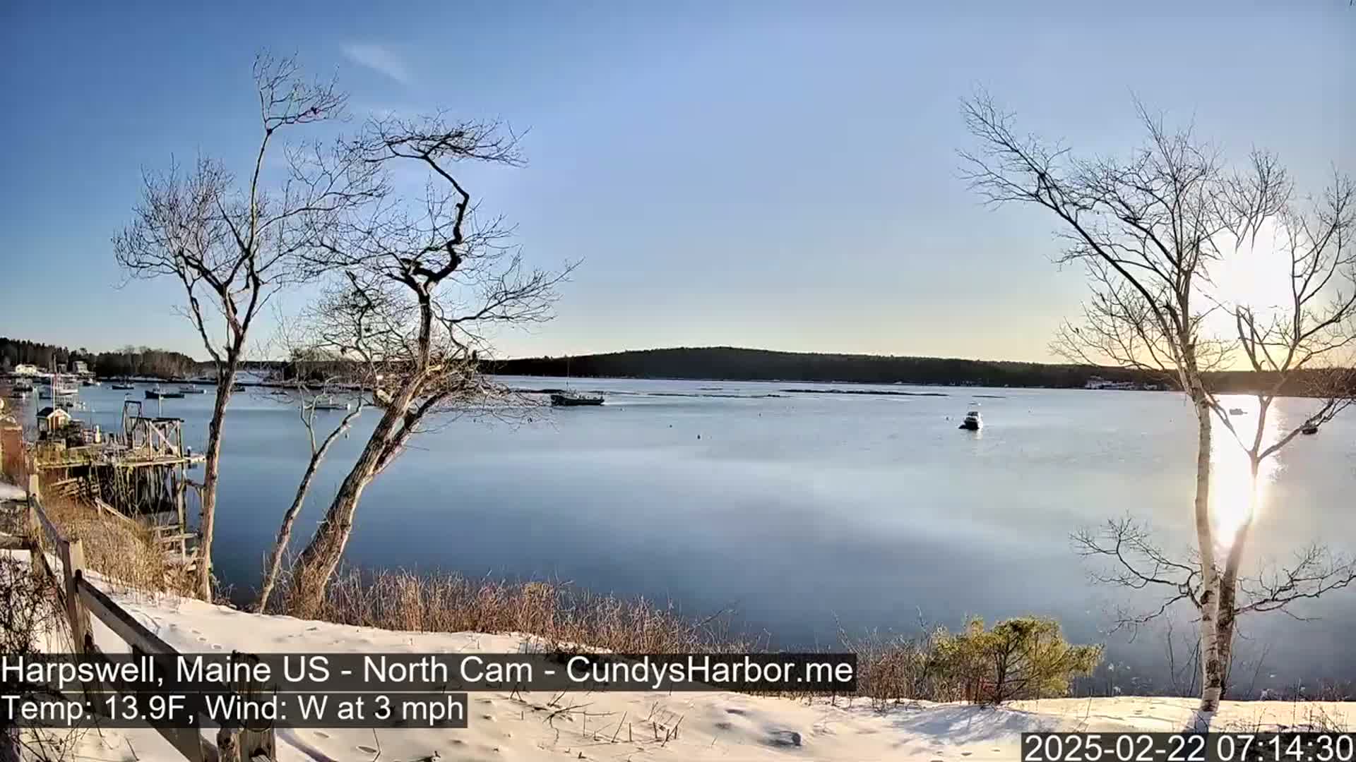 A snow-covered shoreline with bare trees overlooks a calm, partially frozen harbor with a few boats under a clear, sunny sky.
