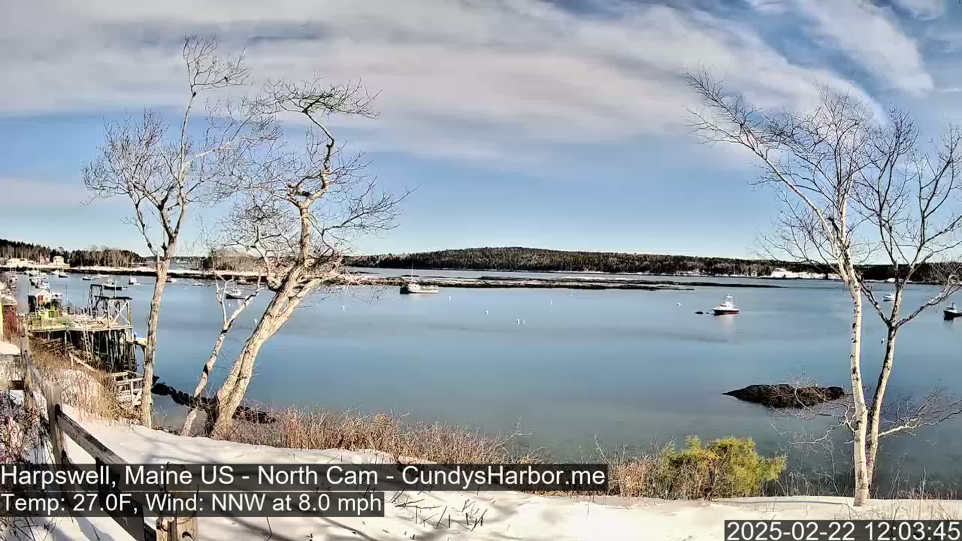 A partly cloudy, cold day with a light north-northwest wind shows a snow-dusted shoreline overlooking a calm bay with several boats, and bare trees in the foreground.