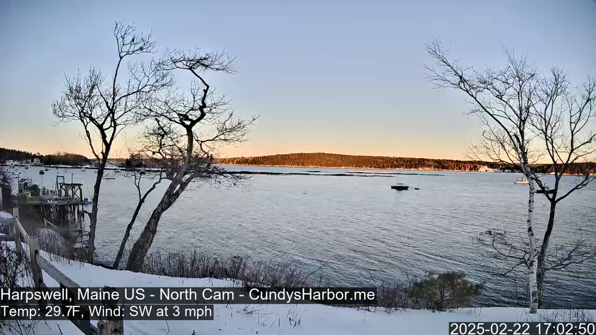 A snow-covered shoreline overlooks a calm body of water with a few boats, under a twilight sky.