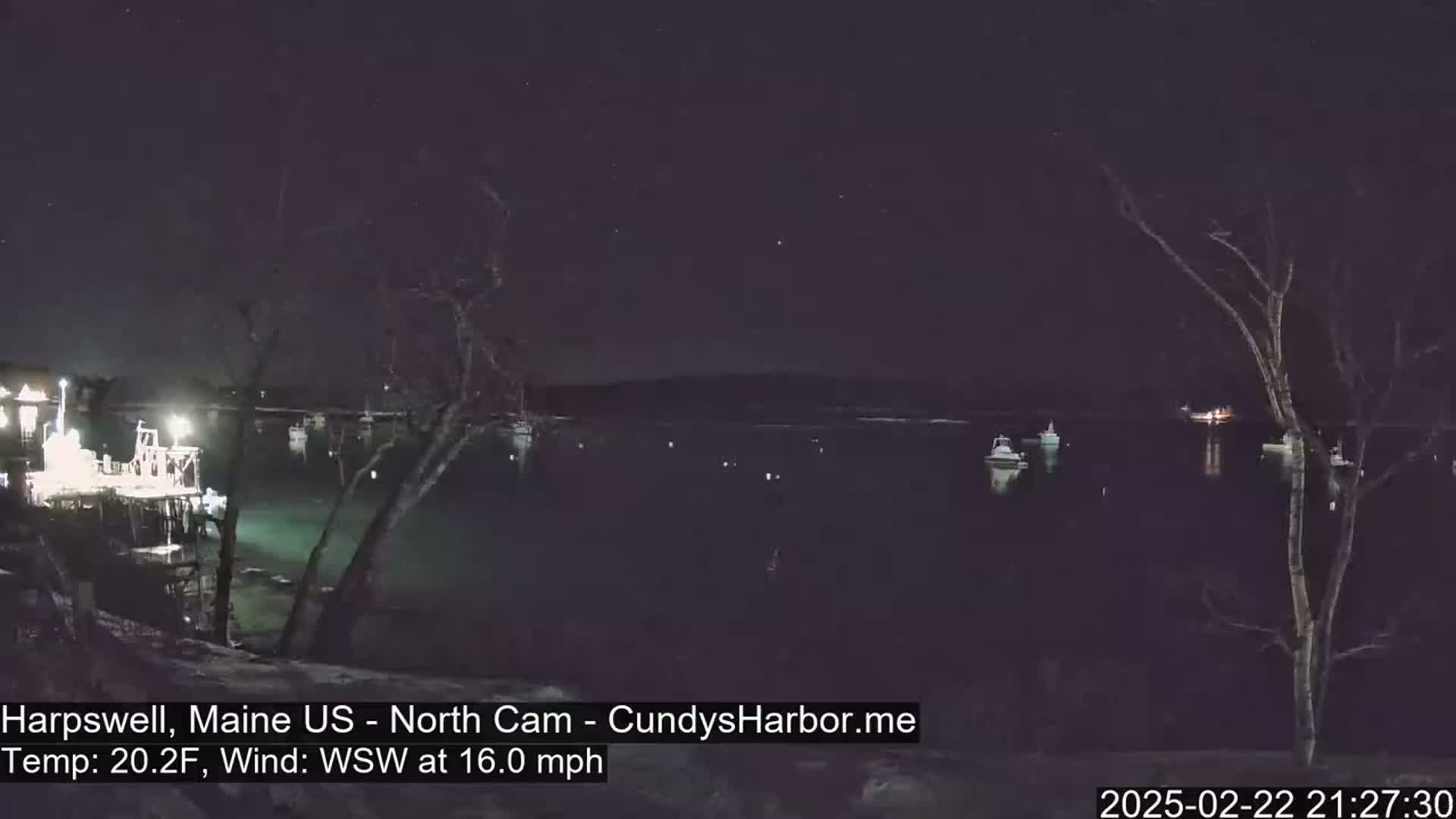 A nighttime view of a dark harbor with several boats, lit faintly, under a clear, starlit sky, and a strong wind.