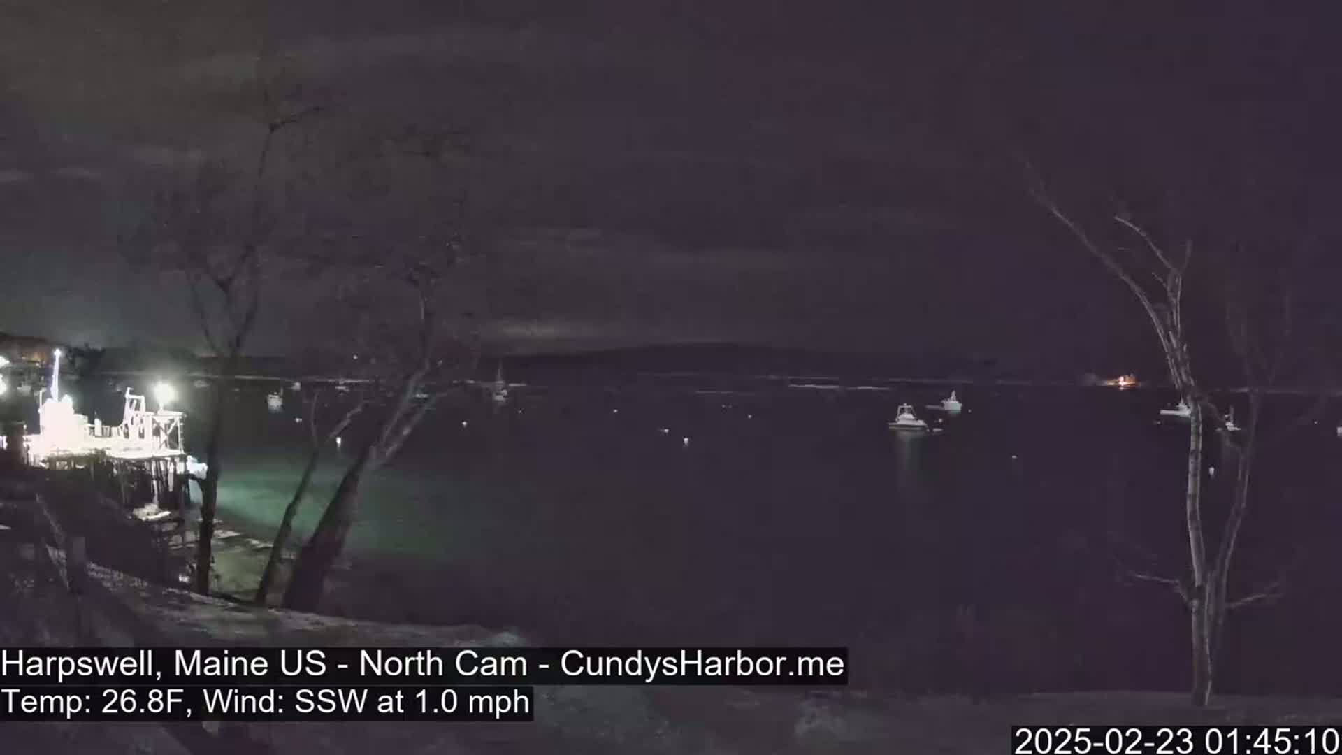 A nighttime view of a harbor with several boats moored, under a dark sky with a slight south-southwesterly wind.
