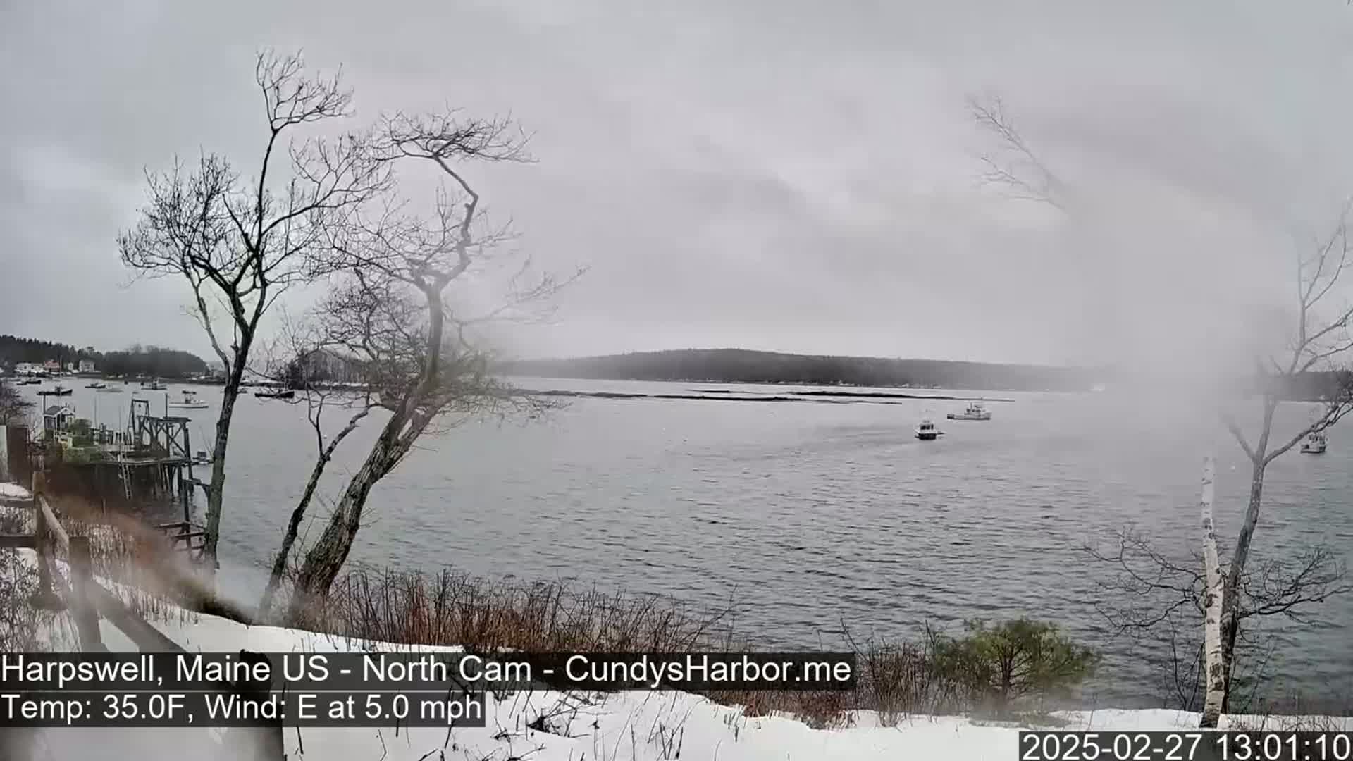 A calm, overcast day with a light east wind shows a snow-covered shoreline overlooking a body of water with several small boats, and bare trees in the foreground.