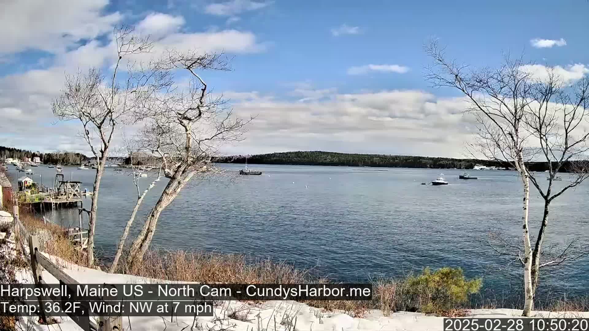 A partly cloudy, cool day with a northwest wind overlooks a calm bay with several boats and a snow-dusted shoreline.