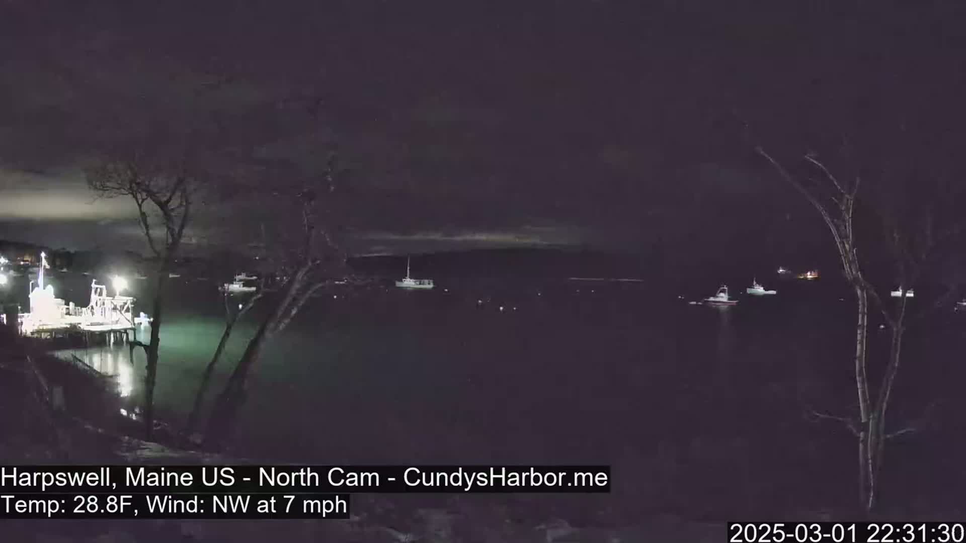 A nighttime harbor scene with several boats at rest under a dark, cloudy sky, a northwest wind blowing at 7 mph, and a temperature of 28.8 degrees Fahrenheit.