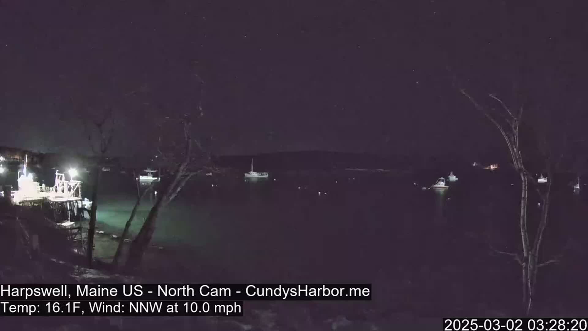A nighttime view of a harbor with several boats moored, under a starry sky, with a 10 mph northwest wind and a temperature of 16.1 degrees Fahrenheit.