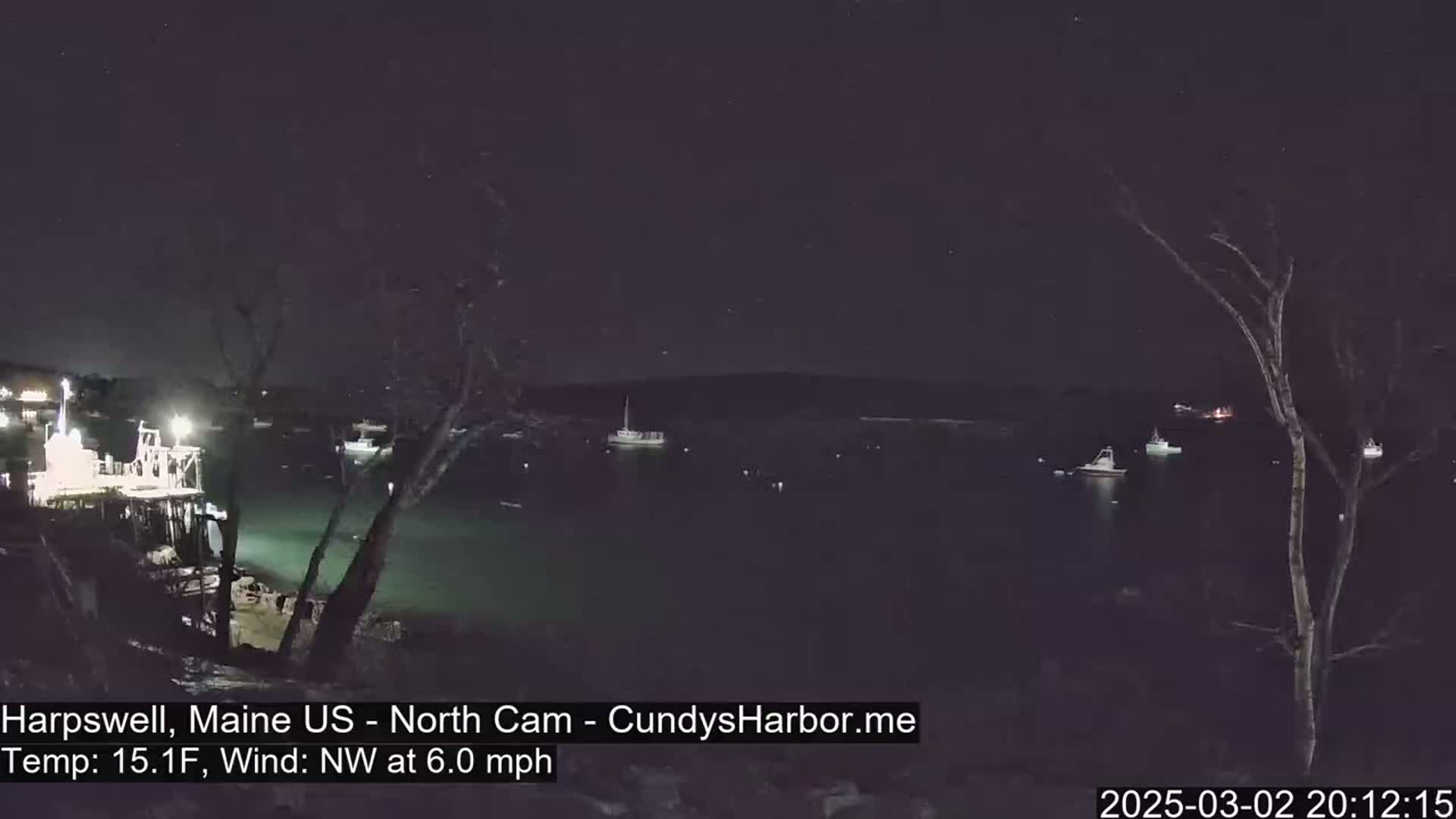 A nighttime view of a harbor with several boats, under a clear, starry sky, with a northwest wind at 6.0 mph and a temperature of 15.1°F.