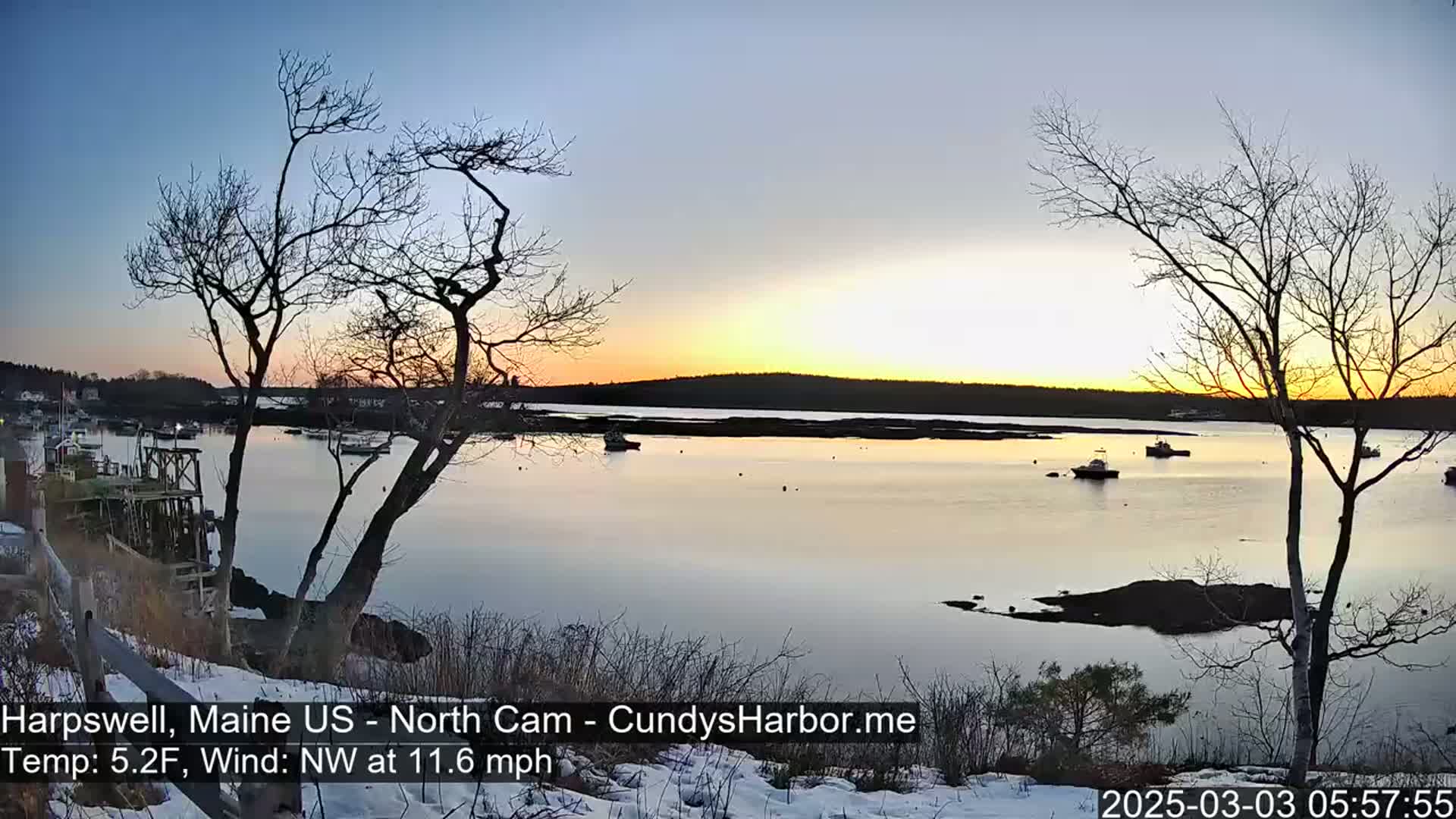 A calm bay at sunset, with a few boats and bare trees in the foreground, under a clear sky and light northwest wind, and some snow on the ground.