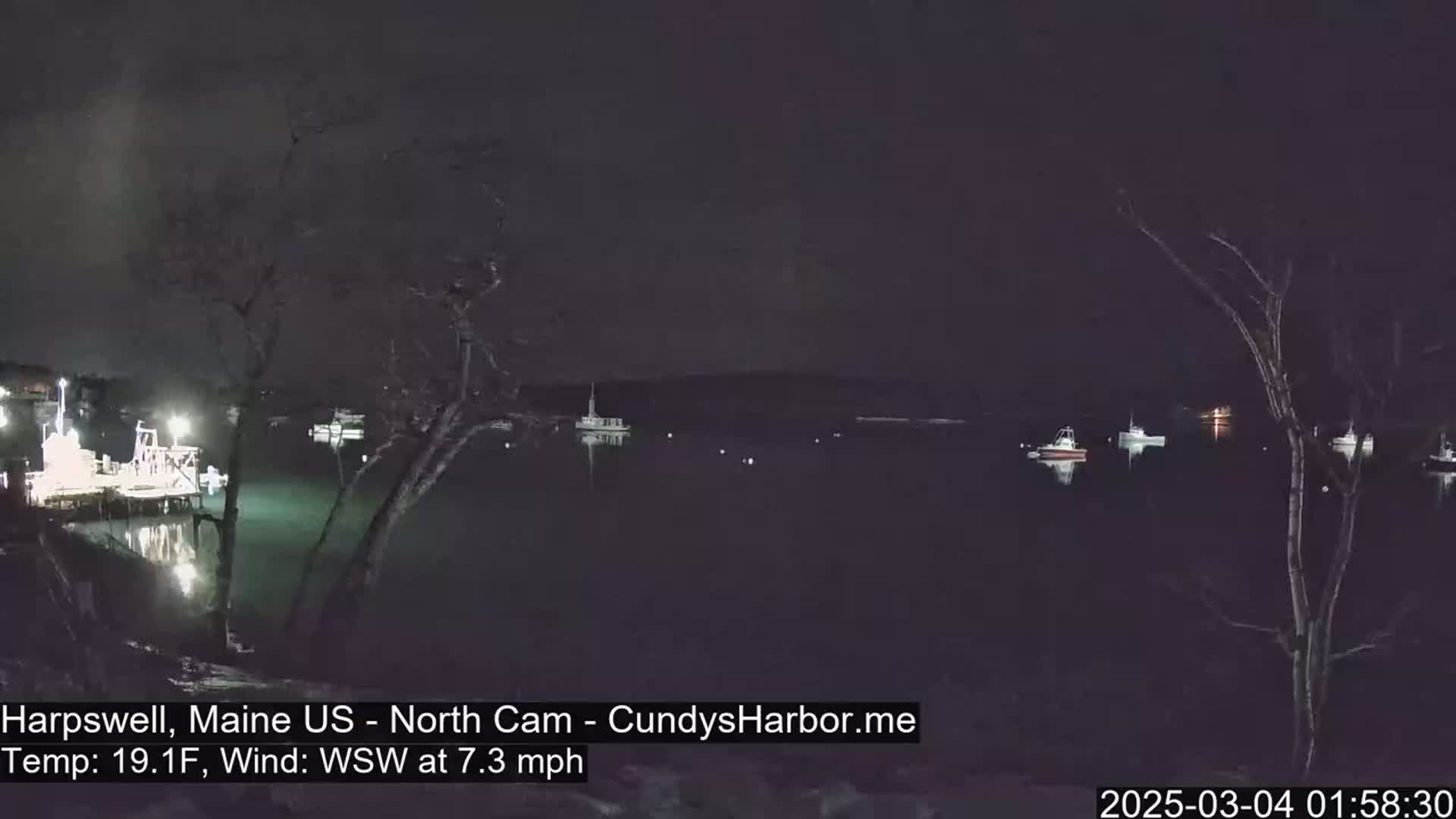 A nighttime view of a calm harbor with several boats moored, under a dark sky with a 19.1°F temperature and a 7.3 mph wind from the west-southwest.