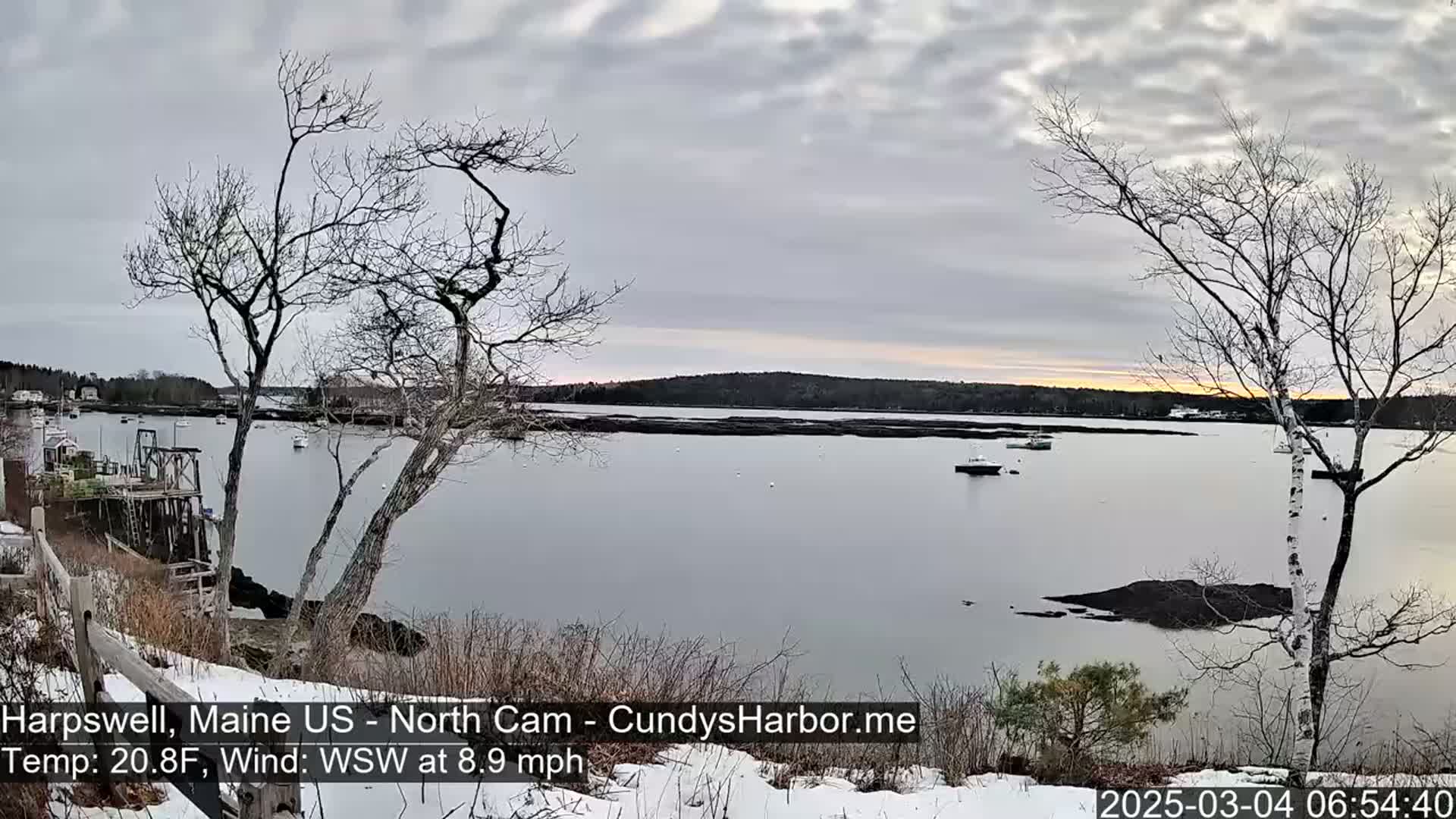 A calm, gray-sky day with a few boats on a body of water, viewed from a snow-dusted shoreline with leafless trees in the foreground.