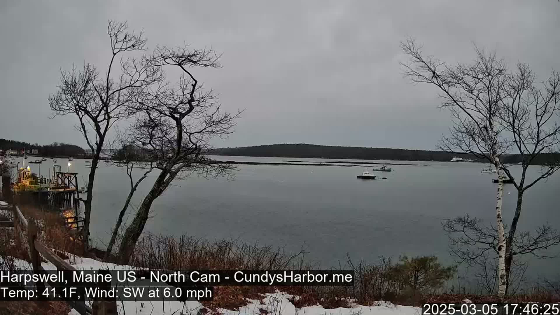 A calm, gray day with a southwest wind shows a view of a harbor with several boats, a dock, and bare trees dusted with snow in the foreground.