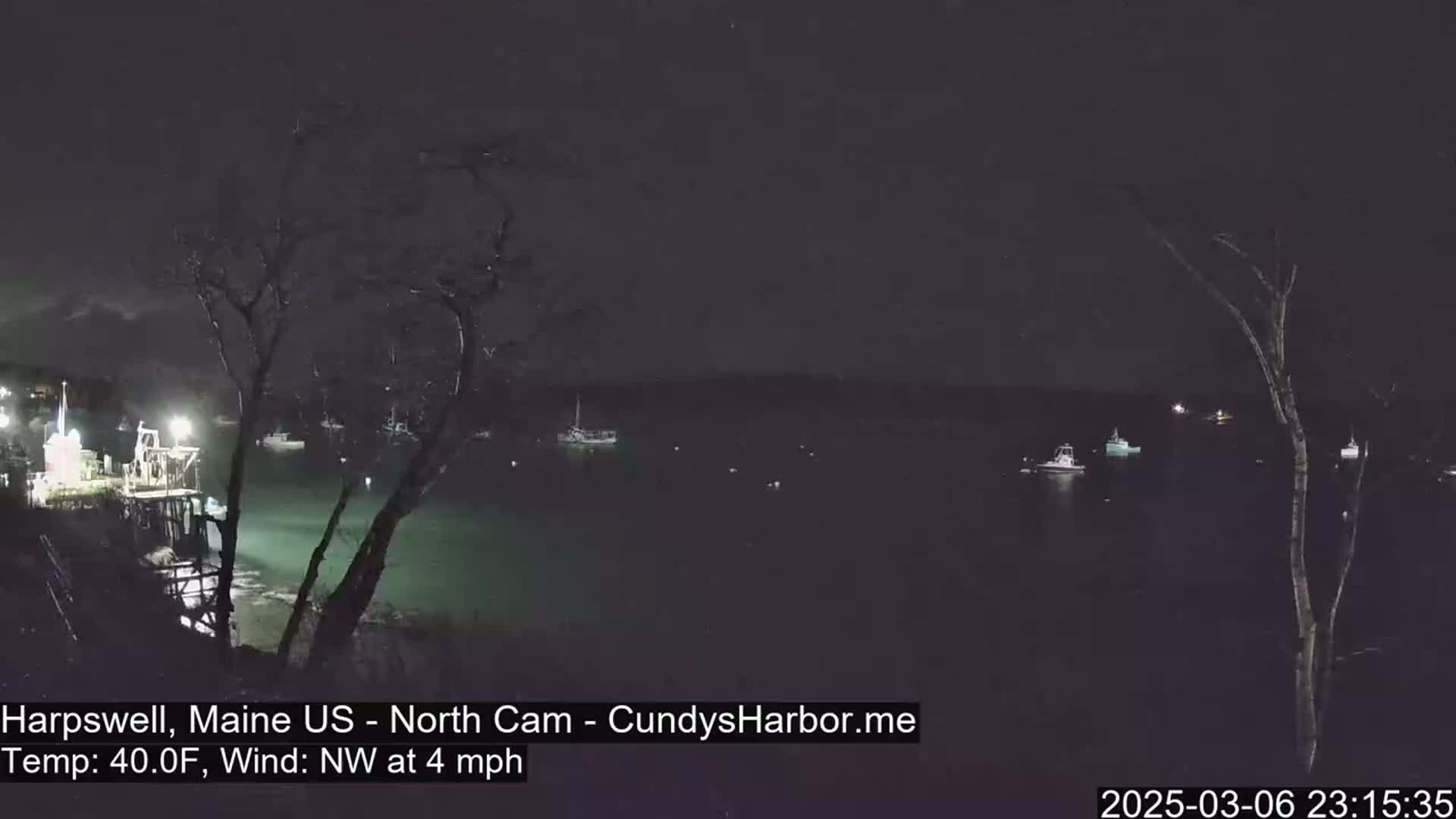 A nighttime view of a harbor with several boats, lit by faint lights, under a dark sky with a northwest wind at 4 mph and a temperature of 40°F.