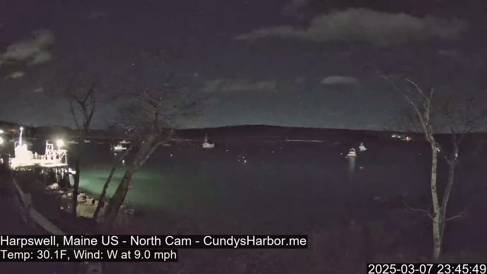 A nighttime view of a harbor with several boats moored, under a partly cloudy sky with a 9 mph westerly wind and a temperature of 30.1°F.