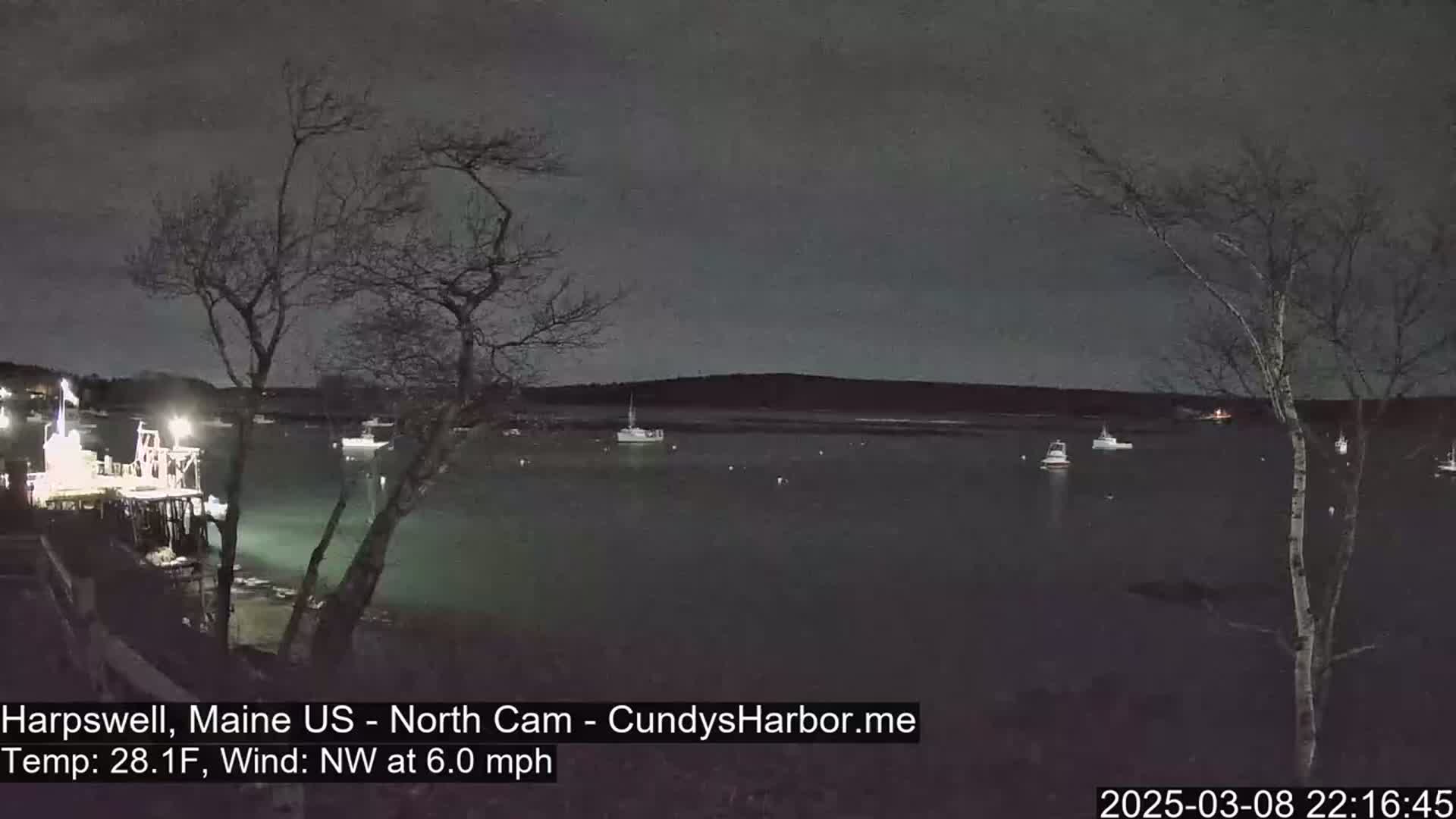 A nighttime view of a calm harbor with several boats moored, under a dark, cloudy sky.