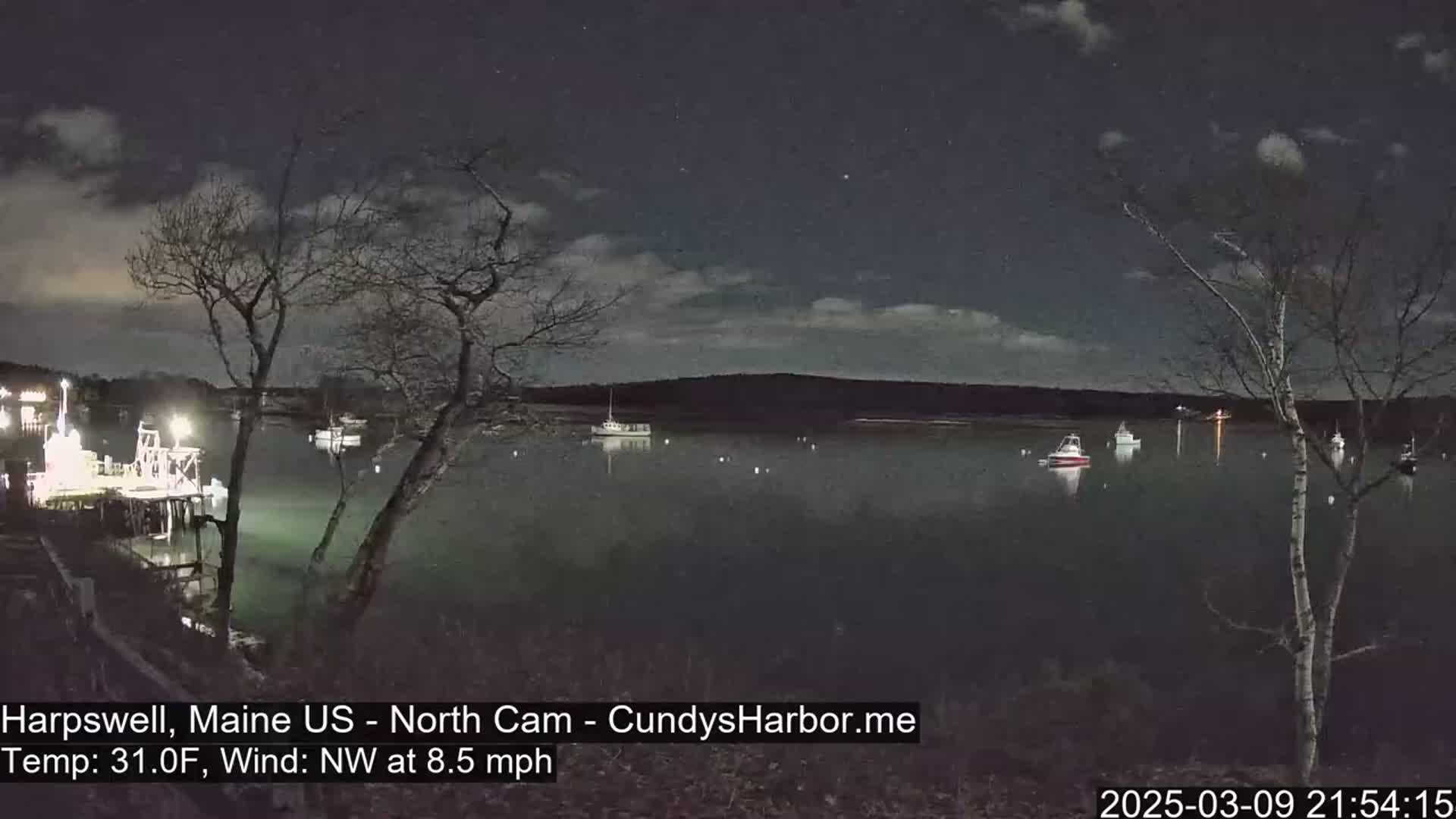 A nighttime view of a calm harbor with several boats moored, under a partly cloudy sky and a northwest wind of 8.5 mph, with the temperature at 31 degrees Fahrenheit.