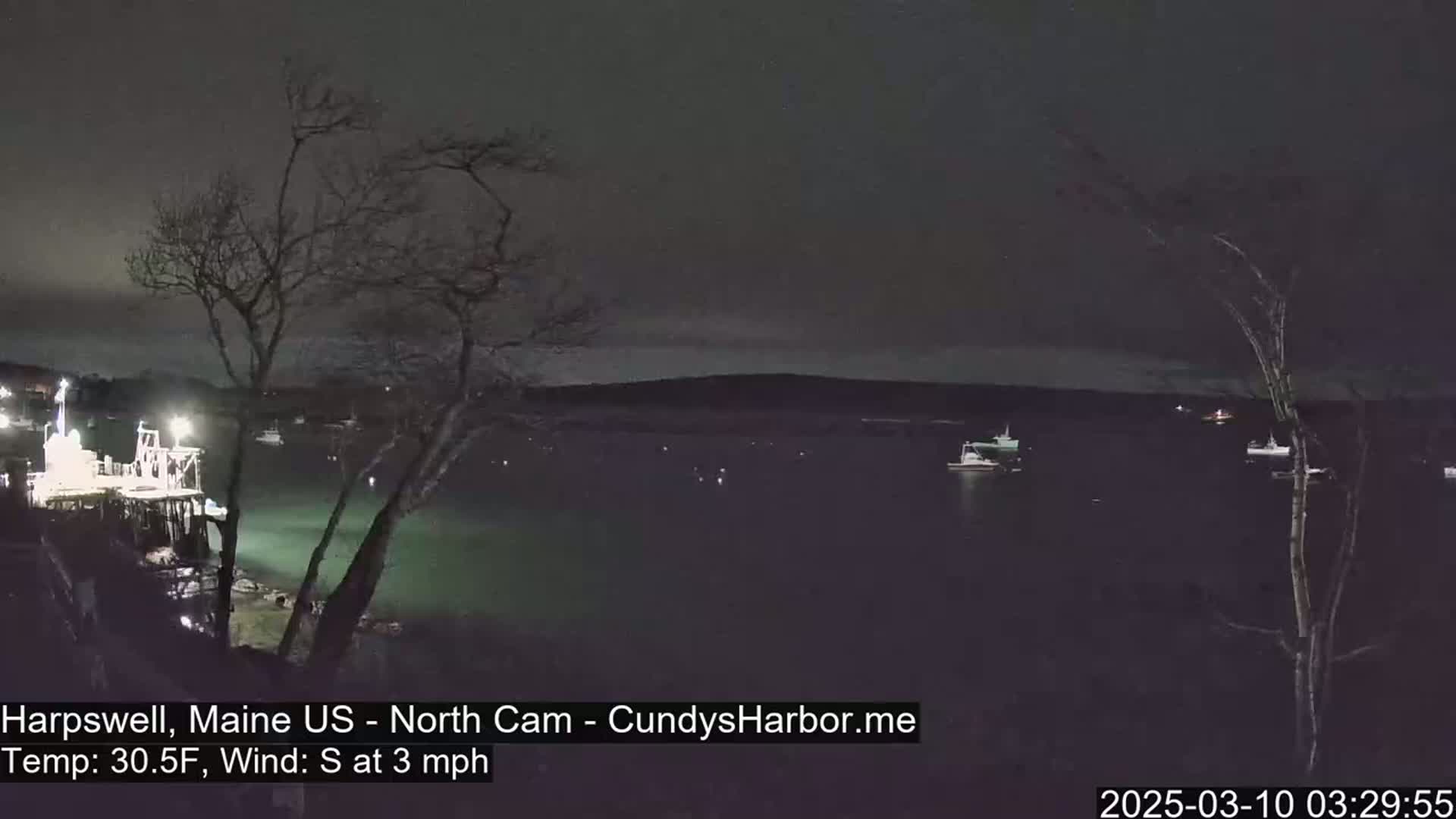 A nighttime view of a dark body of water with several boats moored, under a cloudy sky with a gentle south wind.
