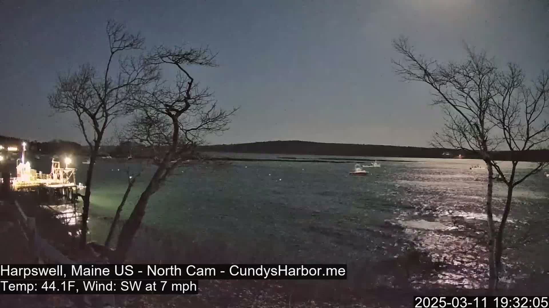 Under a twilight sky, a calm body of water reflects the light, with a few boats and a dock visible near bare trees on the shore, under a southwest wind of 7 mph and a temperature of 44.1°F.