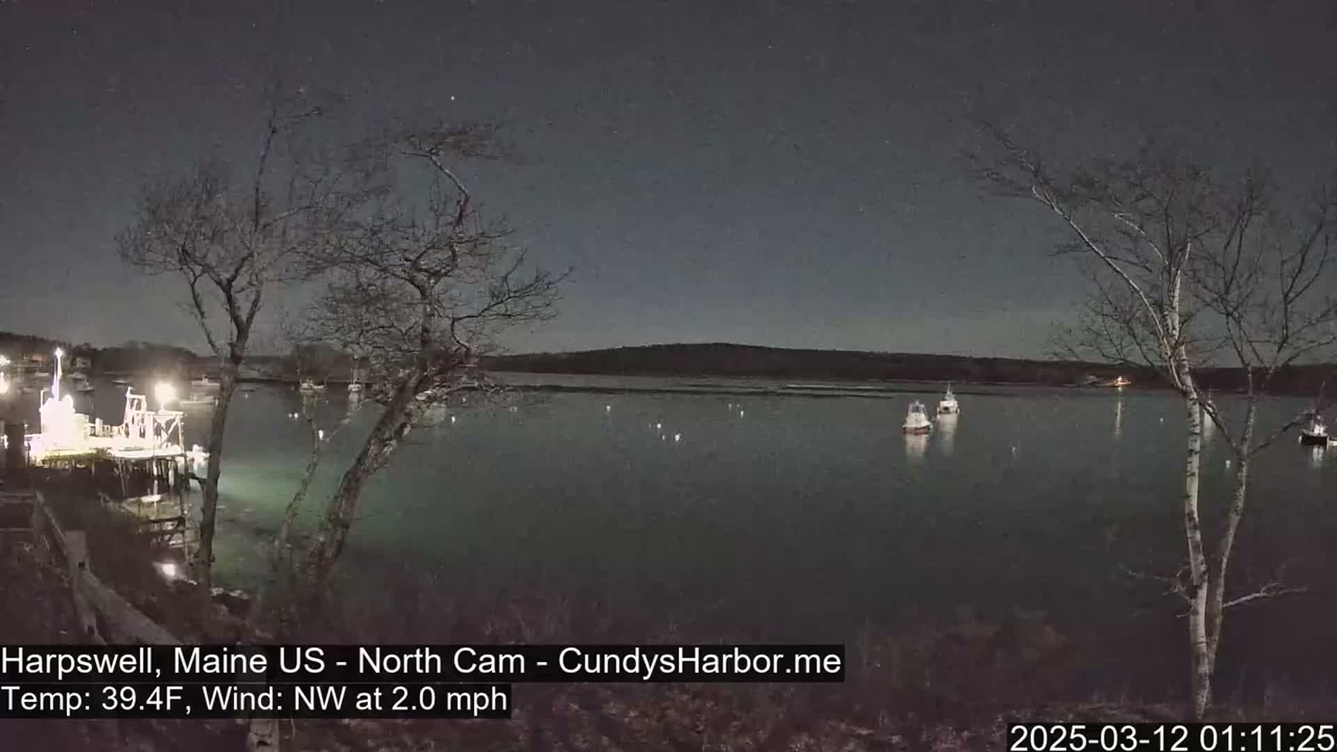 A nighttime view of a calm harbor with several boats moored, under a clear, starry sky, with a gentle northwest wind.