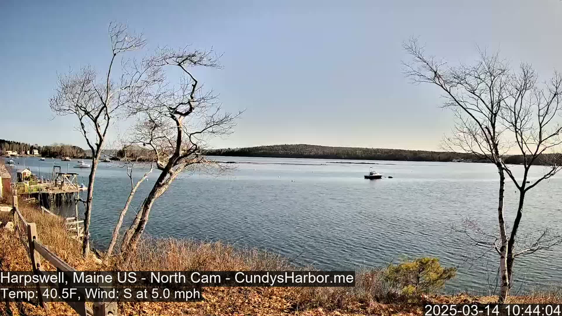 A calm, sunny day shows a view of a harbor with a few boats, some bare trees, and a distant shoreline.