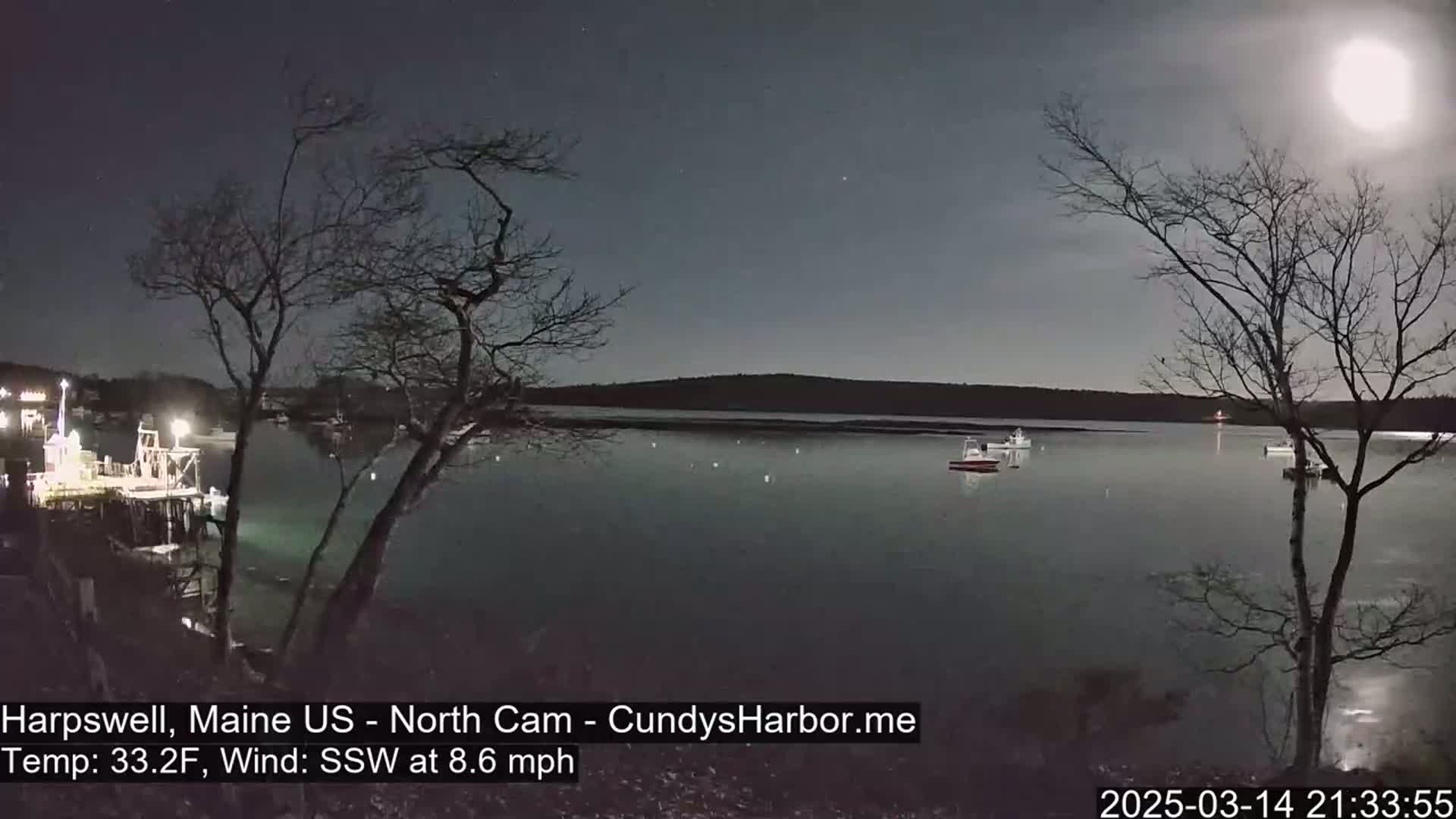 Under a bright moonlit sky with a temperature of 33.2°F and a south-southwest wind at 8.6 mph, several boats are moored in a calm harbor near a dark shoreline with bare trees.