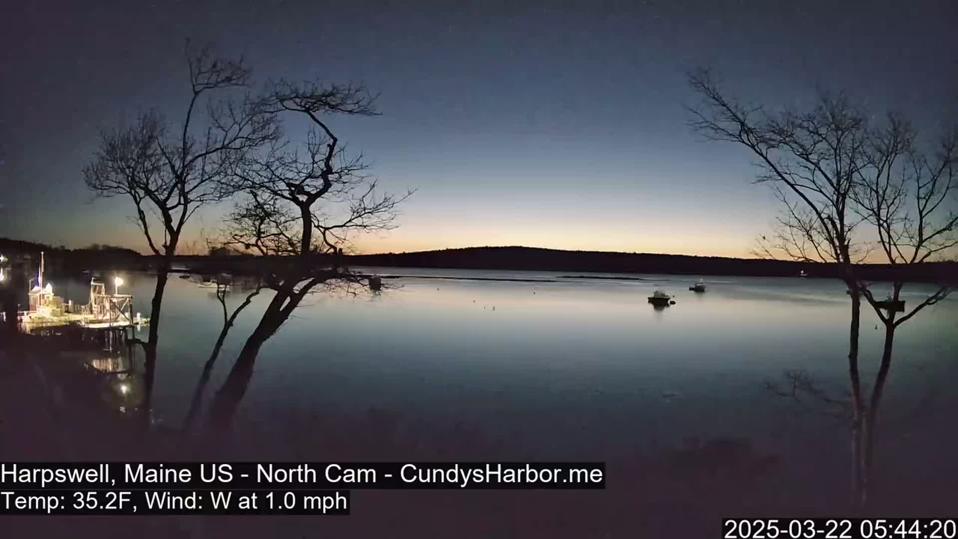 A tranquil, pre-dawn scene shows a calm body of water with a few boats, framed by bare trees under a twilight sky.