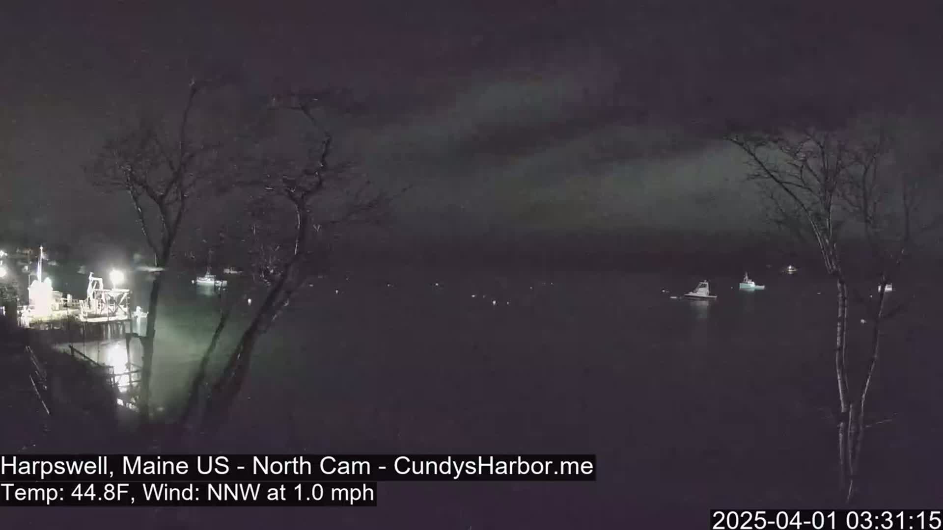 A nighttime view of a harbor with several boats moored, under a dark, cloudy sky with a  north-northwest wind.