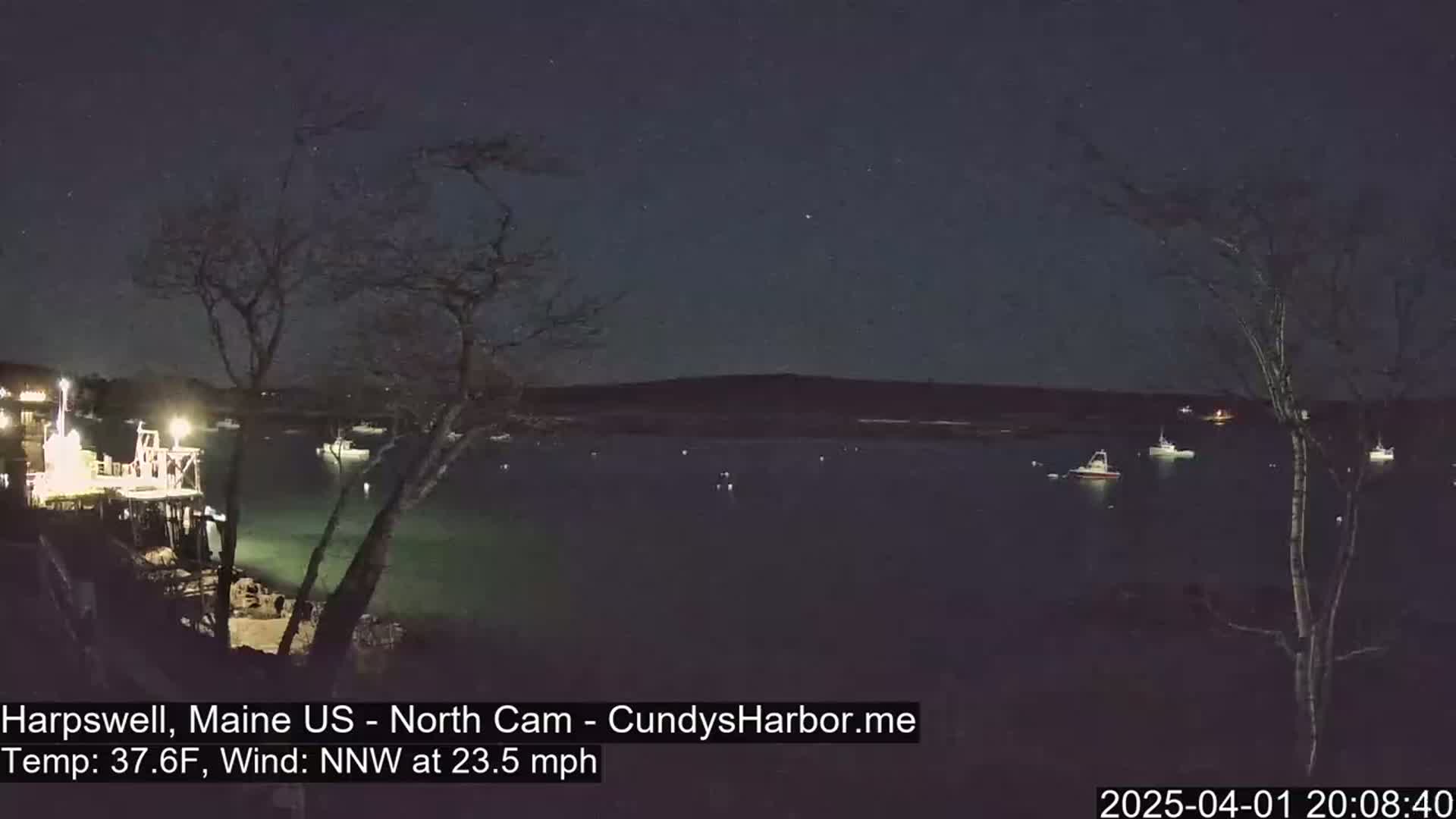 Several boats are moored in a dark body of water under a clear, starry night sky with a 23.5 mph northwest wind and a temperature of 37.6°F.