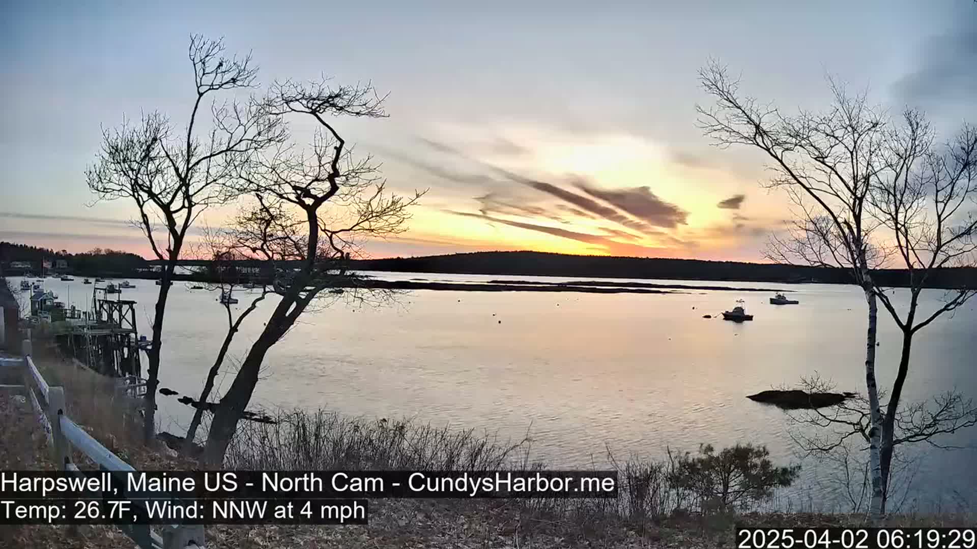 A calm body of water with several boats is seen at sunset under a partly cloudy sky, with bare trees in the foreground.